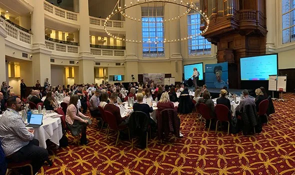 A large conference or seminar in a spacious, elegant ballroom with many attendees seated at round tables, listening to a speaker at a podium on stage, with large screens displaying presentation slides.