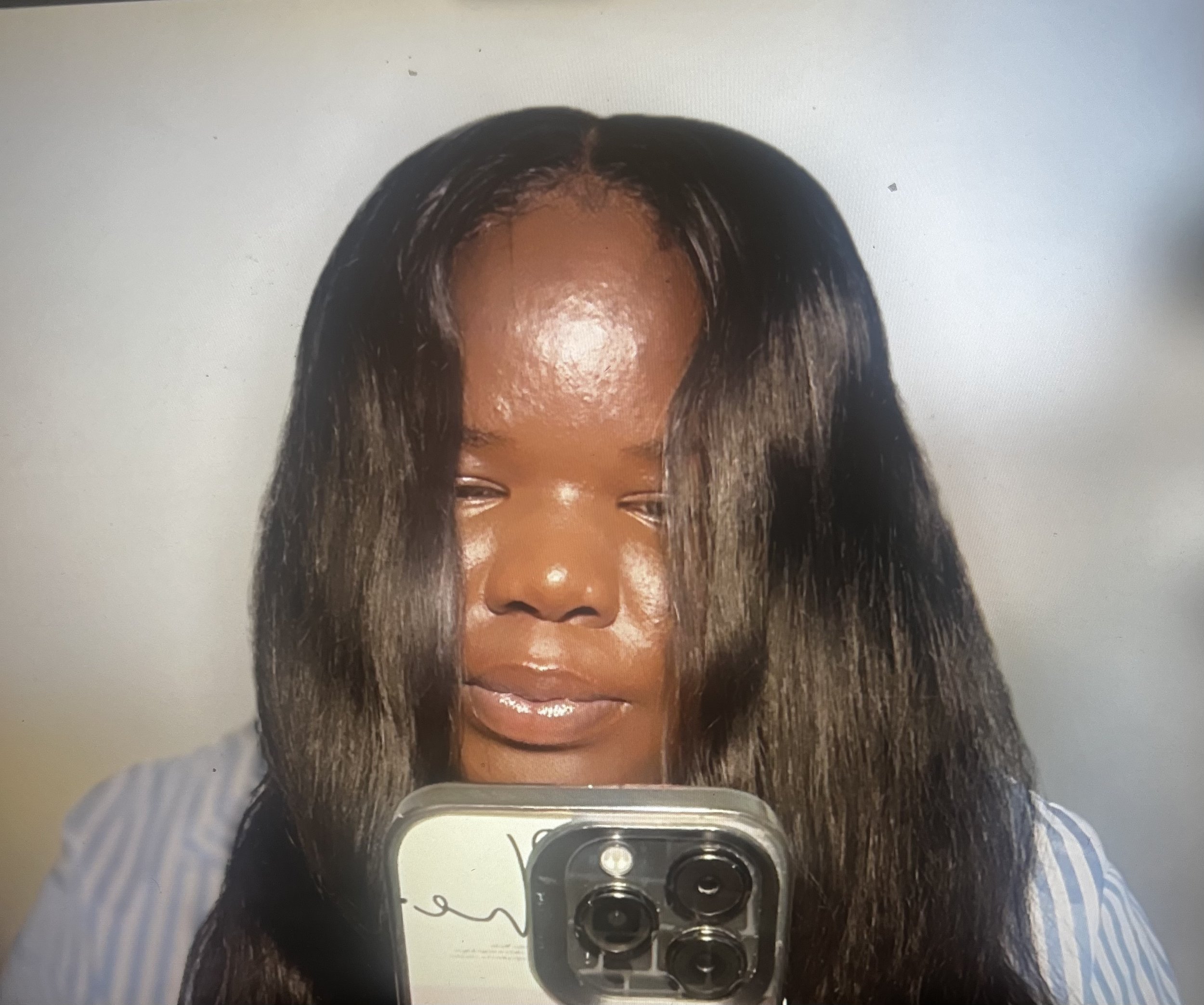 A woman taking a selfie with her phone. Her face is partially visible, and she has long, dark hair. She appears to be smiling, with a white top and striped shirt visible. The background is neutral and plain.