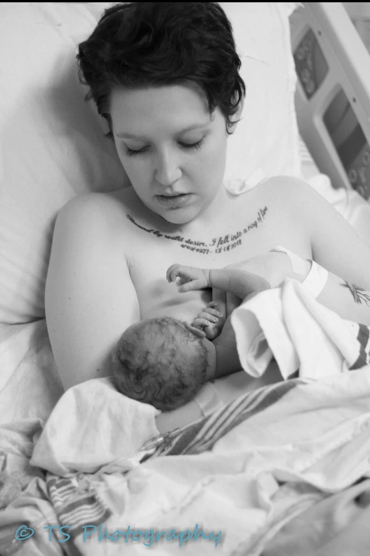 Black and white photograph of a woman with short dark hair breastfeeding a newborn in a hospital bed, with a tattoo on her chest and a hospital monitor in the background.
