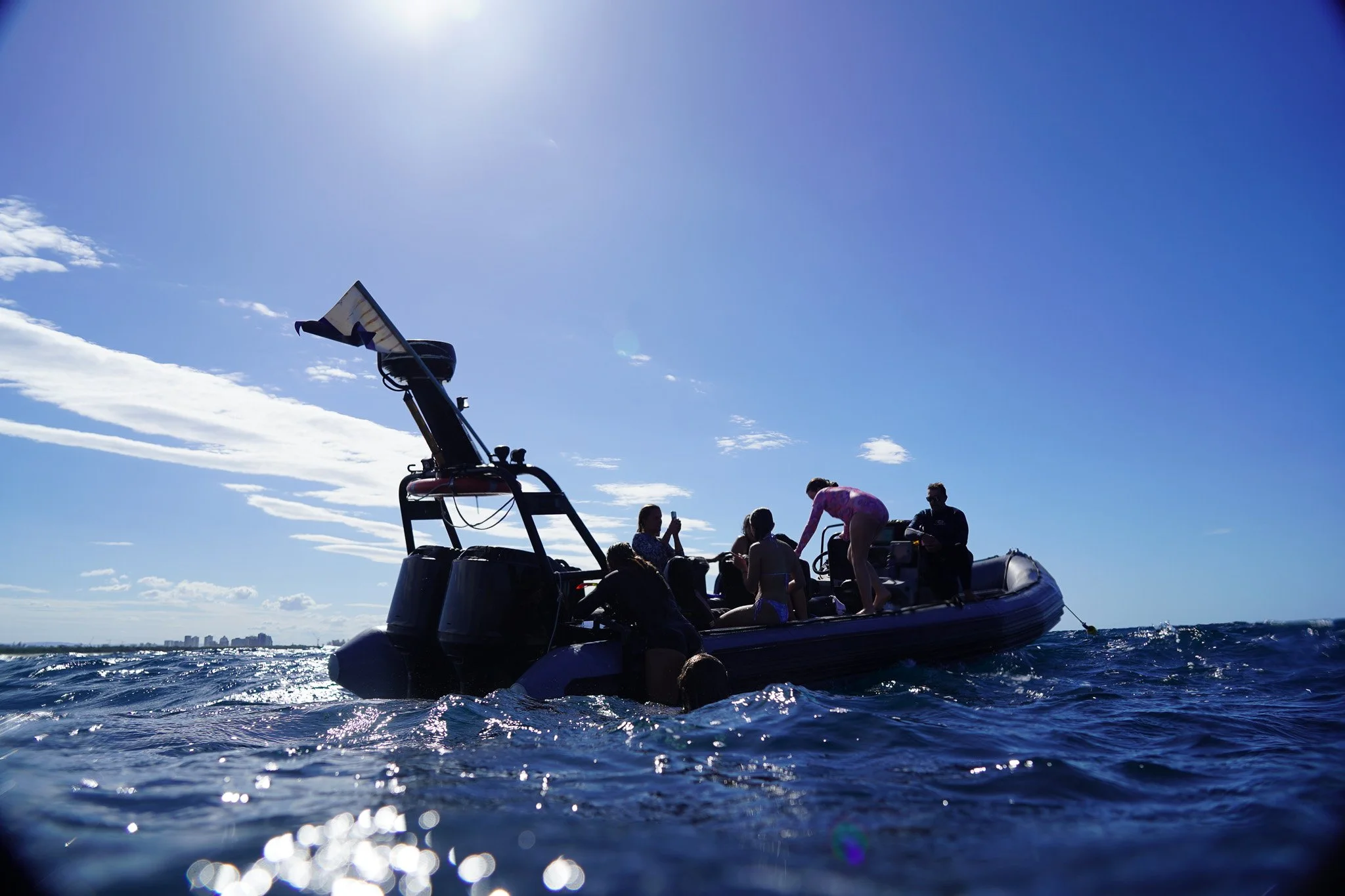 Group of people on a boat in the ocean under a bright blue sky, some sitting and some standing, with a city skyline visible in the distance.