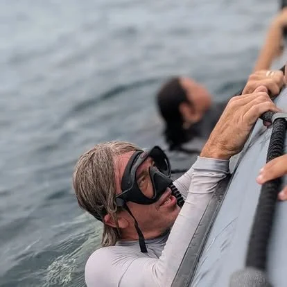 A woman in a wetsuit, wearing a diving mask, holds onto the side of a boat while in the water. Another person is visible in the background.