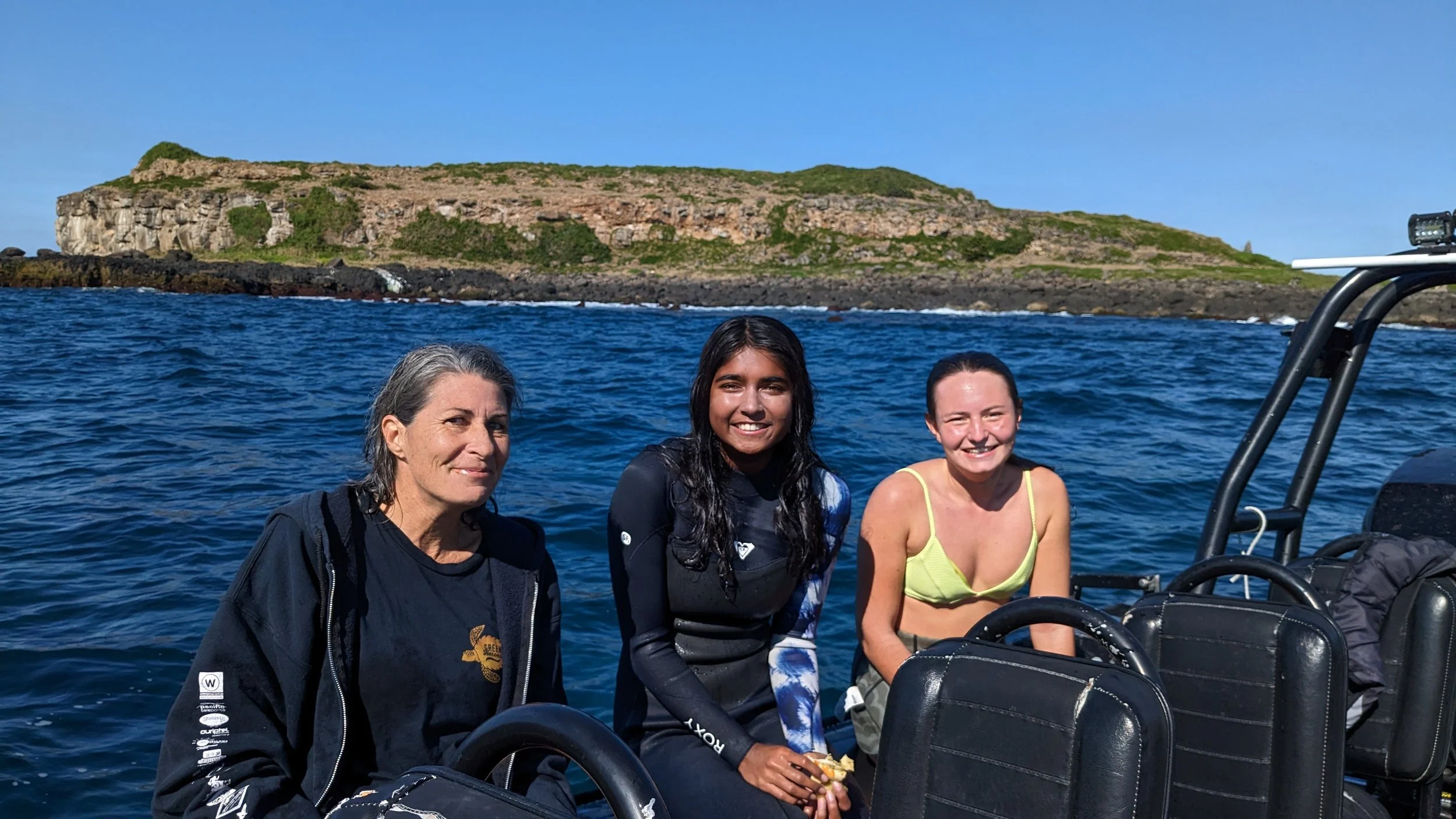 Three women sitting on a boat with ocean and land in the background, smiling at the camera.