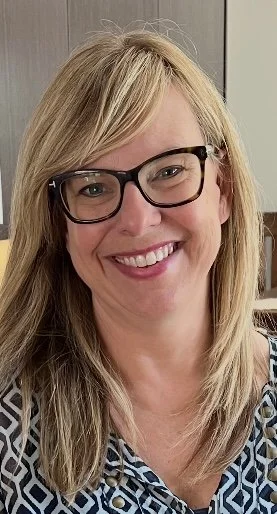 A woman with blonde hair, glasses, and a patterned blouse smiling indoors.