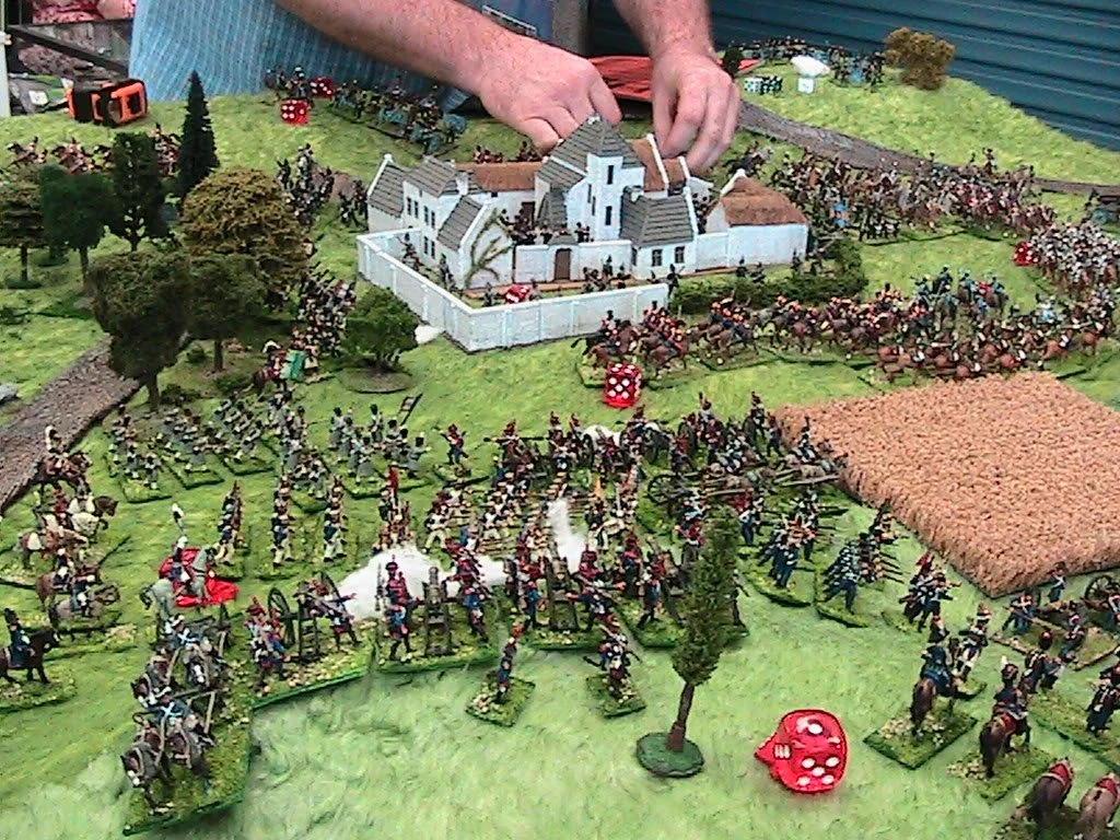 Miniature tabletop battlefield scene featuring toy soldiers, cannons, trees, a white building, and dice, depicting a historical or fictional war.