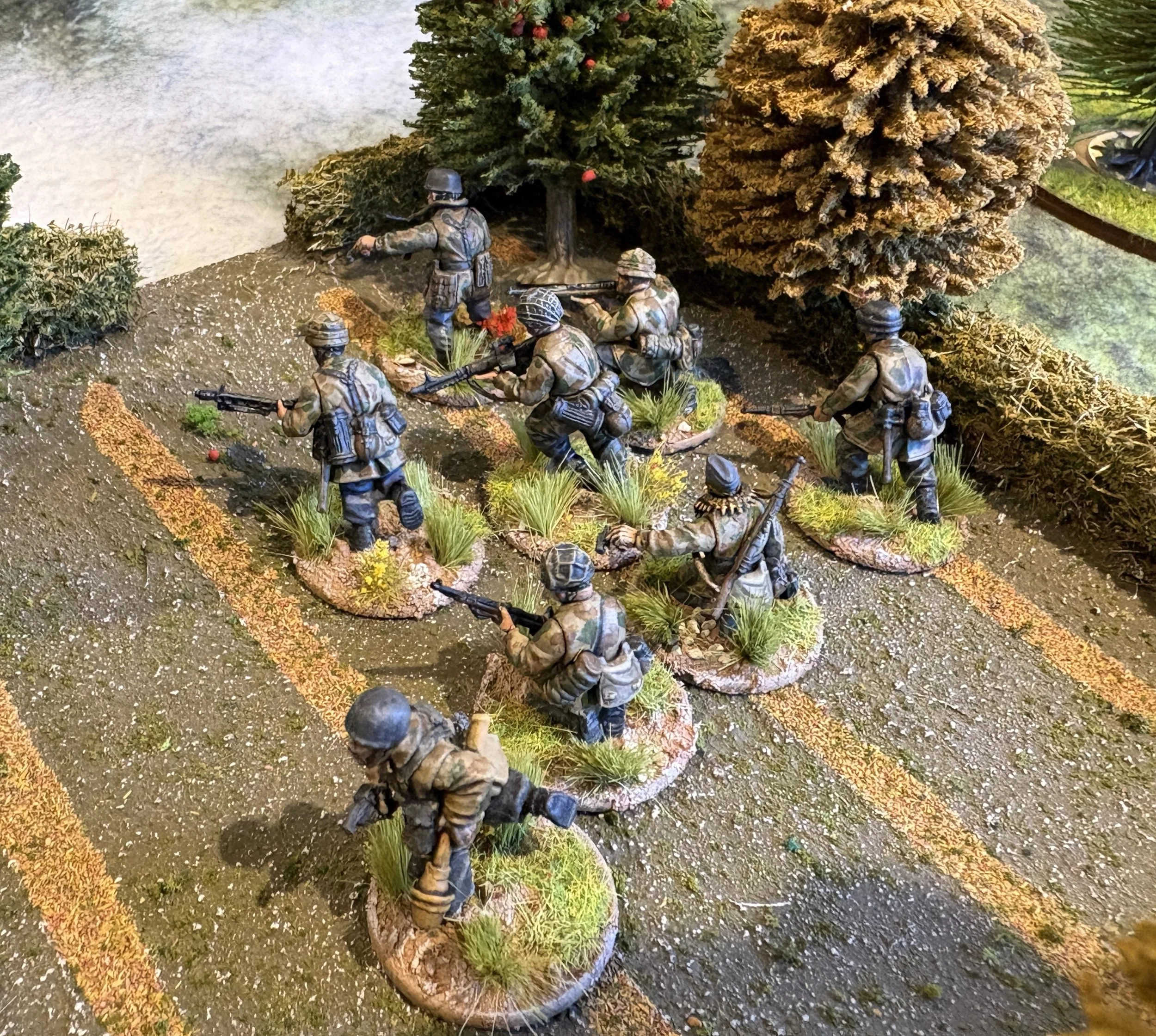 Miniature military figures in camouflage uniforms and helmets, armed with rifles, positioned on a textured terrain with grass and dirt, surrounded by miniature trees and bushes.