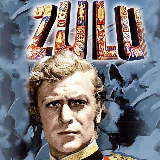 Close-up of a man in a military uniform with a serious expression, standing in front of a background with the word 'ZULU' in colorful, decorative letters.