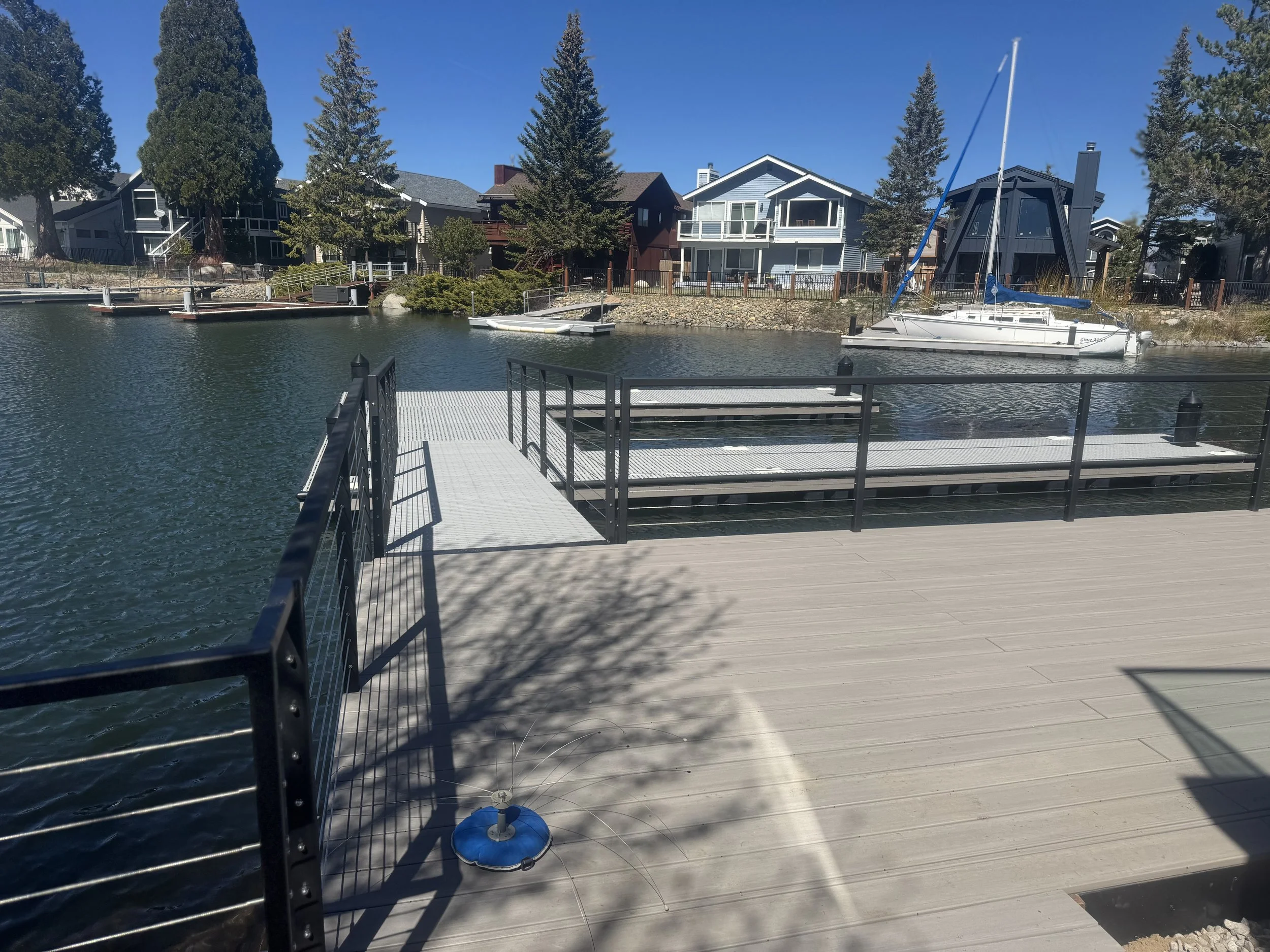A wooden dock with black metal railings extends over water, with a boat tied to the dock in a residential canal lined with modern houses and trees under a clear blue sky.