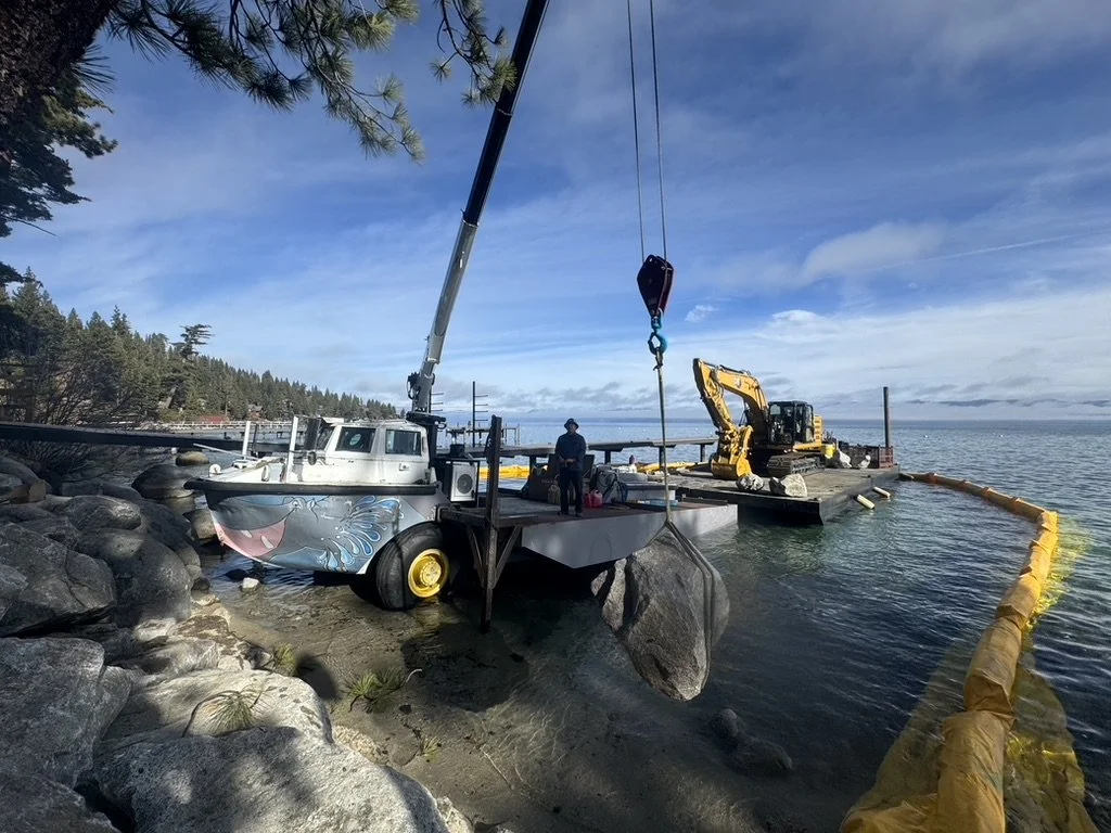 A construction crew is working on a floating platform or barge near a rocky shoreline, using heavy machinery like a crane and small excavator, under a partly cloudy sky.