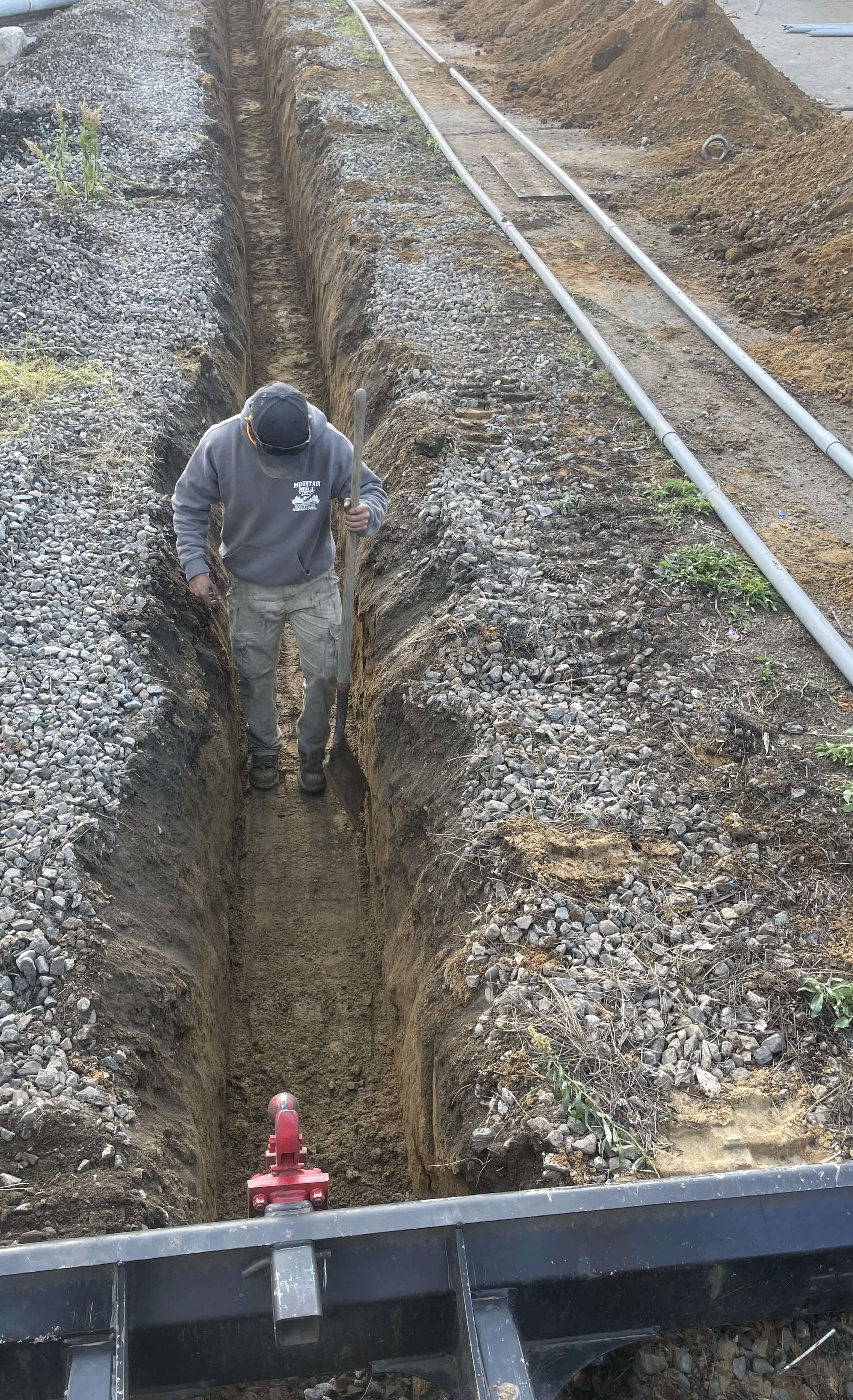 A worker in a gray hoodie and beige pants digging a trench for underground utility pipes or cables on a construction site.