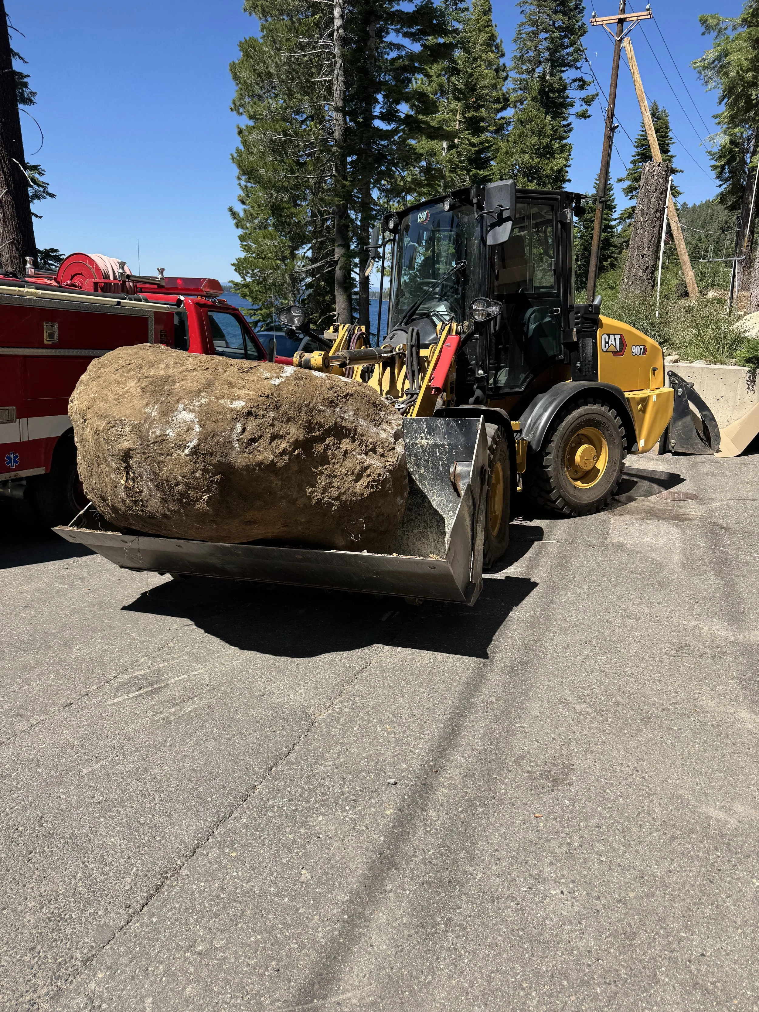 A yellow CAT front loader with a large rock in its bucket, parked on a paved road with a fire truck nearby and tall pine trees in the background.