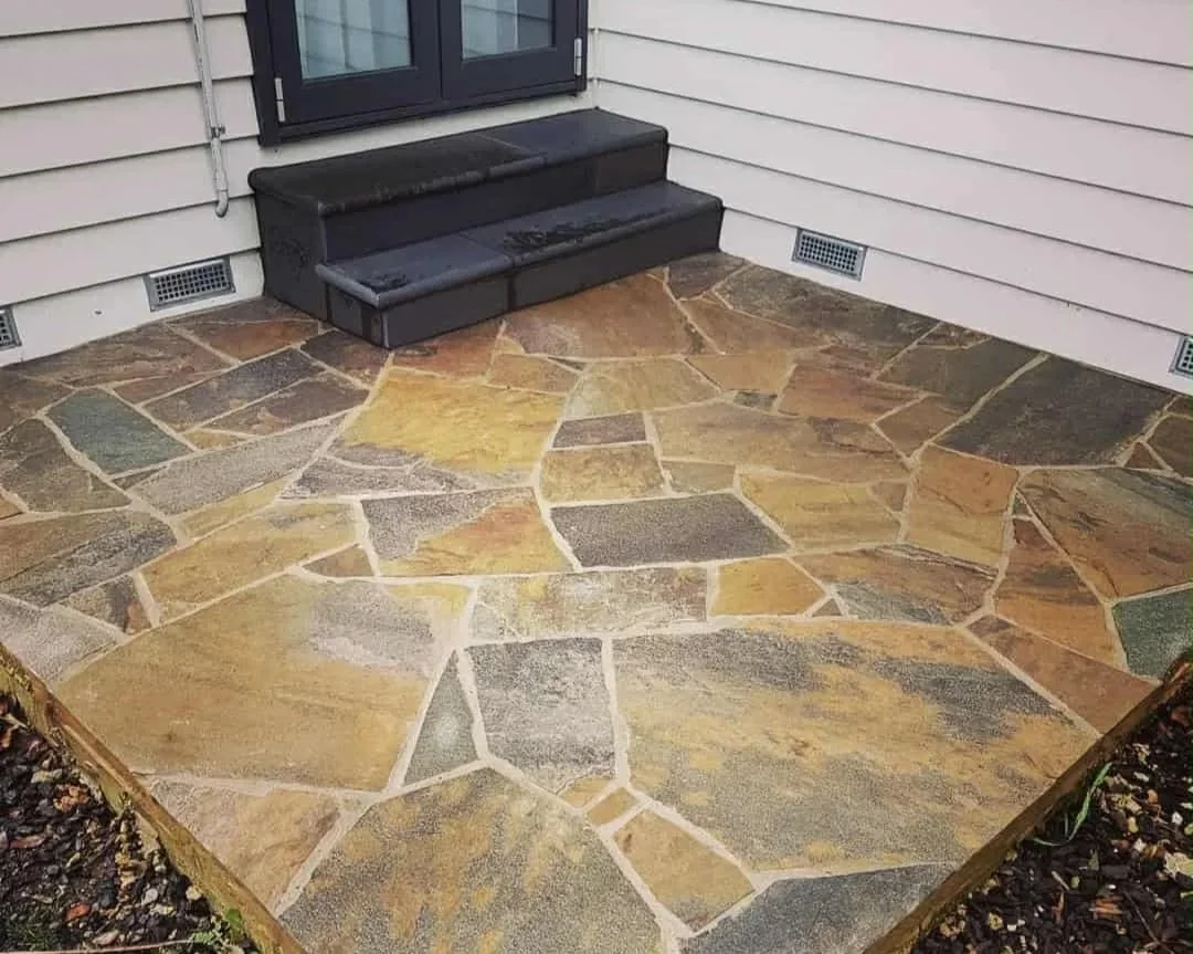 A small outdoor patio with a stone floor and a set of black stairs leading to a door.