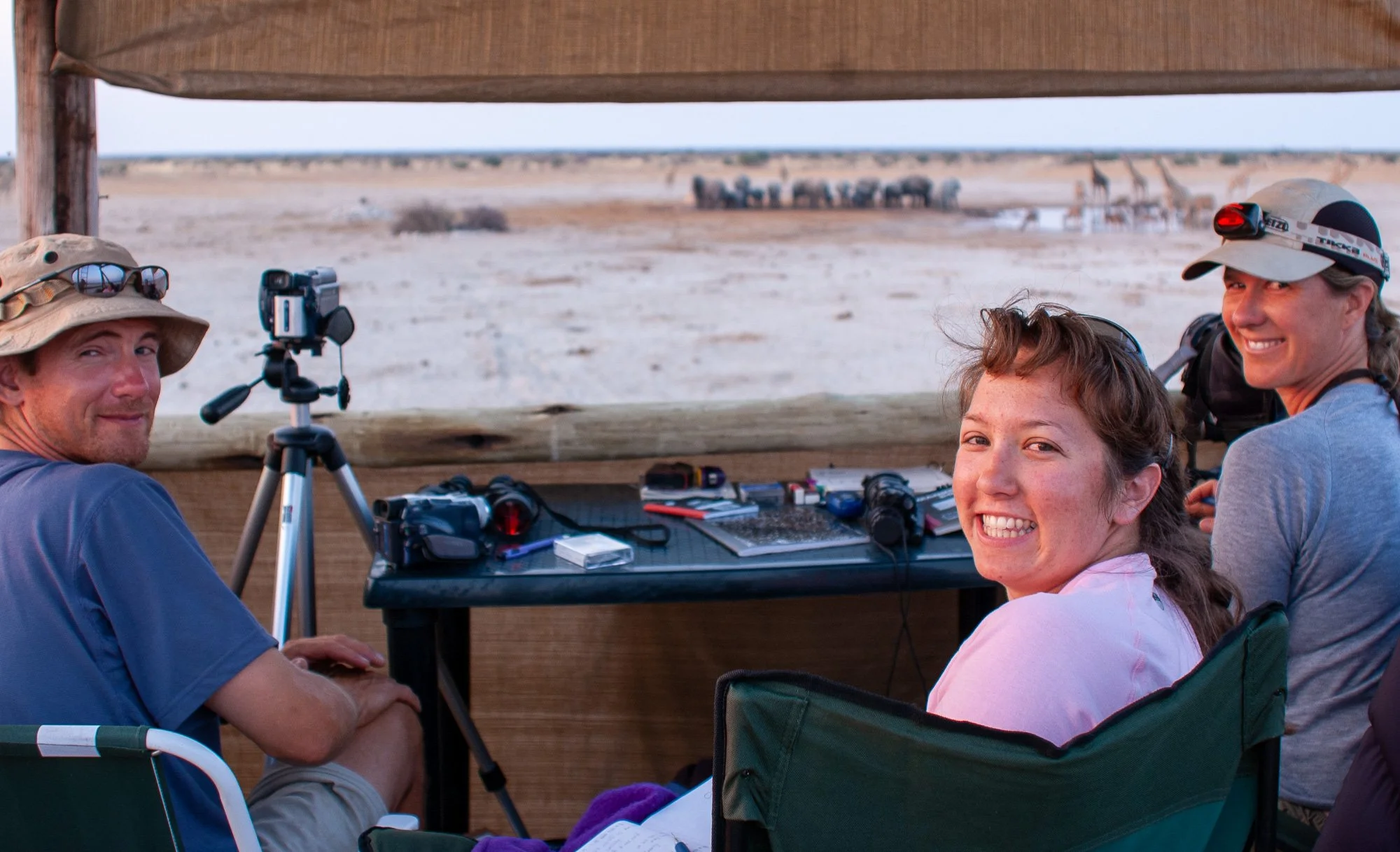 Three people sitting at a table in a safari viewing blind, overlooking an African landscape with giraffes in the distance.