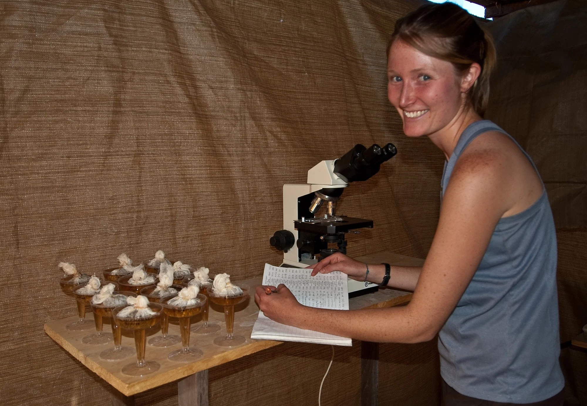 A young woman in a blue sleeveless shirt smiles while looking at a microscope and holding a notebook, standing next to a wooden table with multiple small glasses filled with a dessert topped with cotton candy.