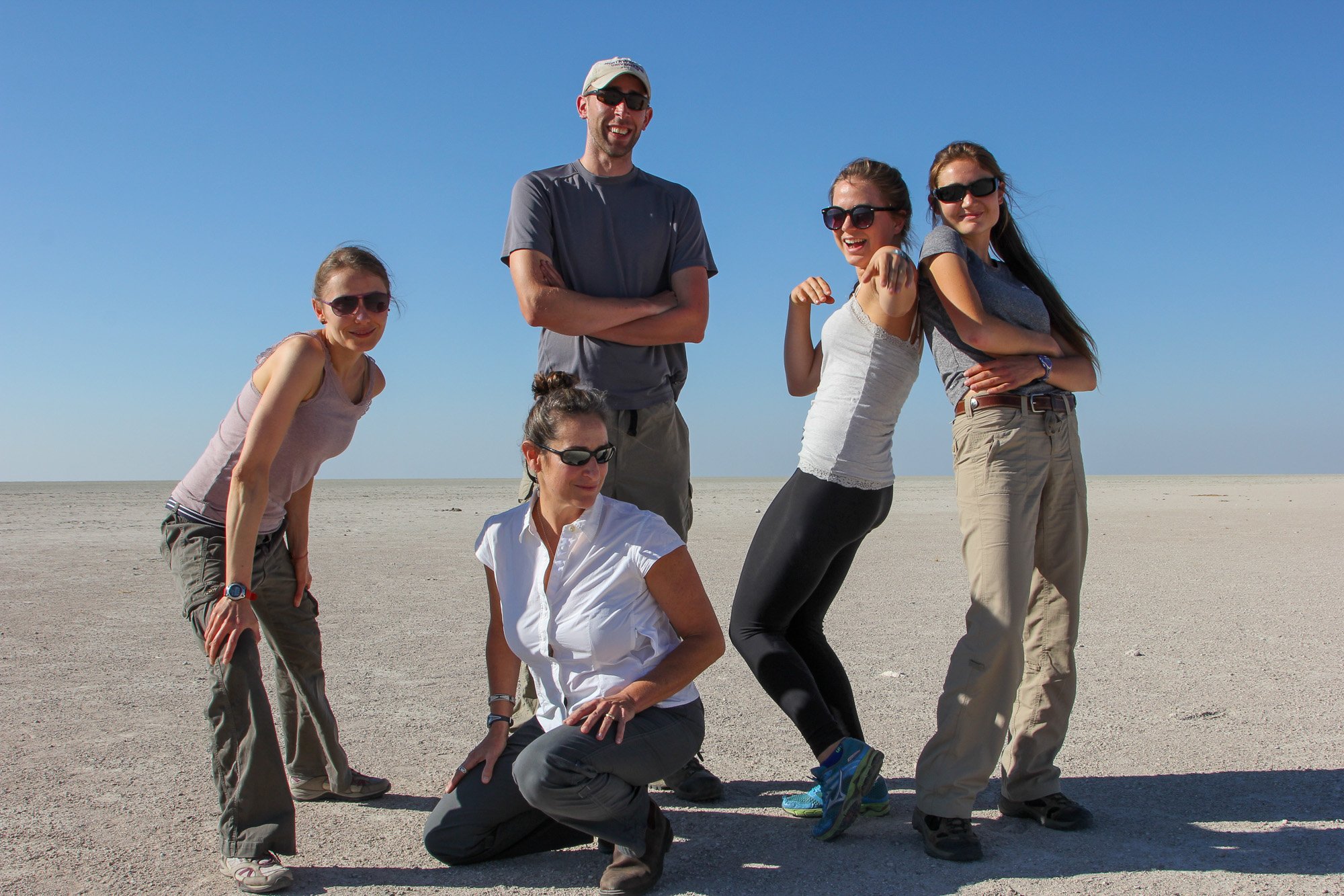 Group of six people in casual outdoor clothes posing on a flat, sandy landscape with clear blue sky.