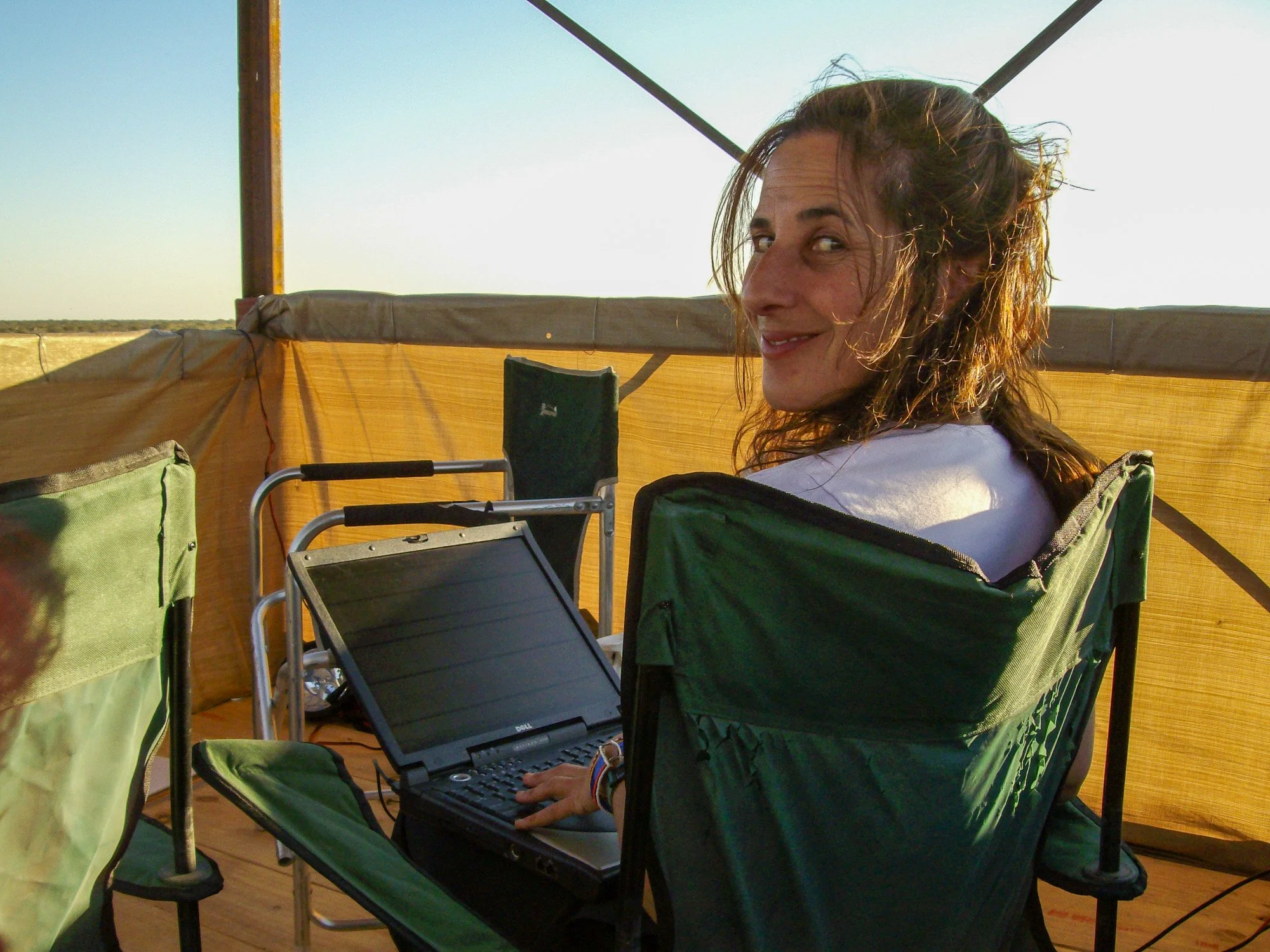 A woman in a white t-shirt sitting in a green camping chair, using a laptop on a small table, outdoors with a yellow privacy screen and blue sky in the background.