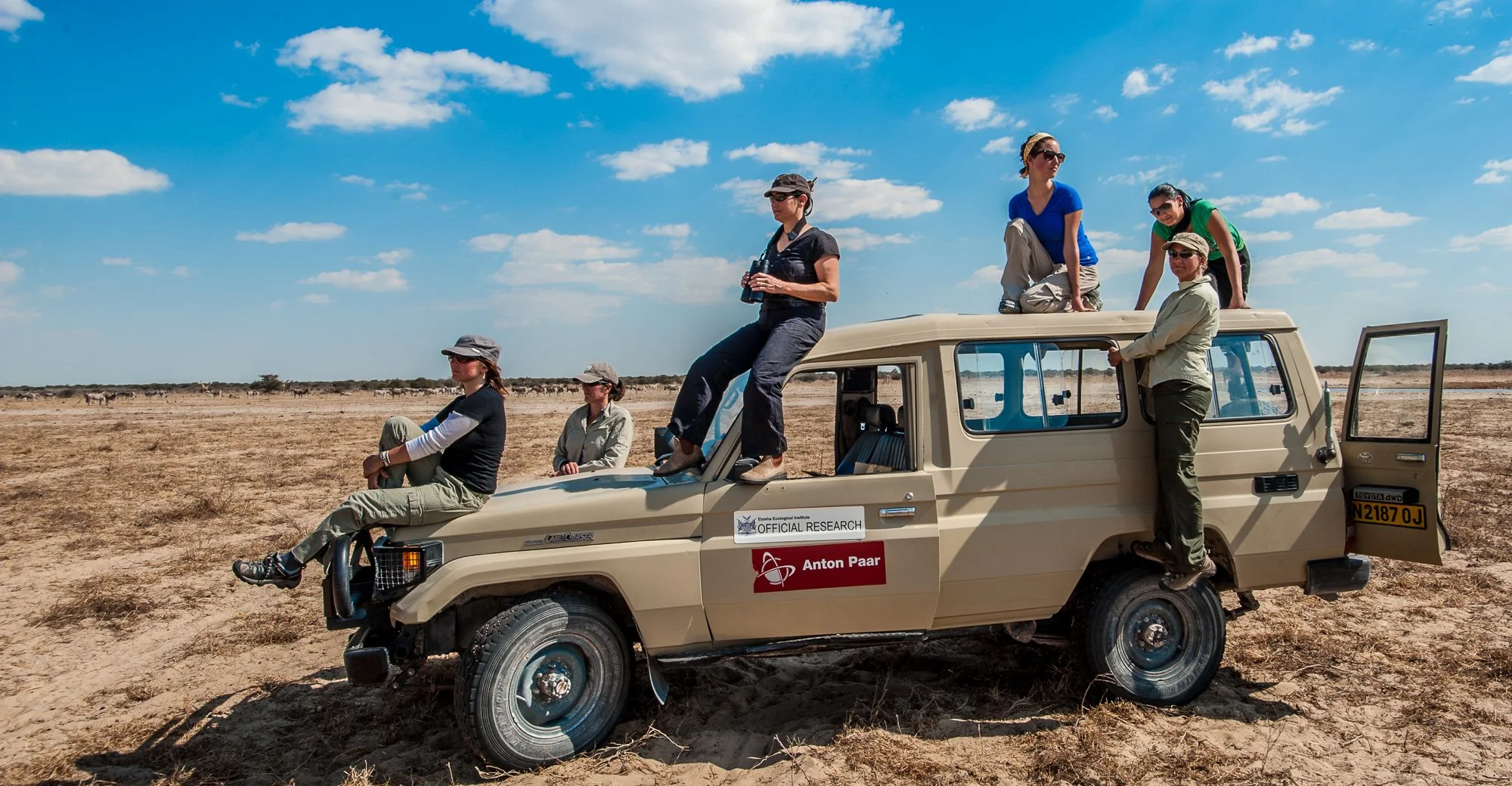 A group of seven women in outdoor clothing and hats sitting or standing on and around a beige Toyota Land Cruiser in a flat, dry, open landscape with a clear blue sky and some clouds. They appear to be on an expedition or research trip, with some loo