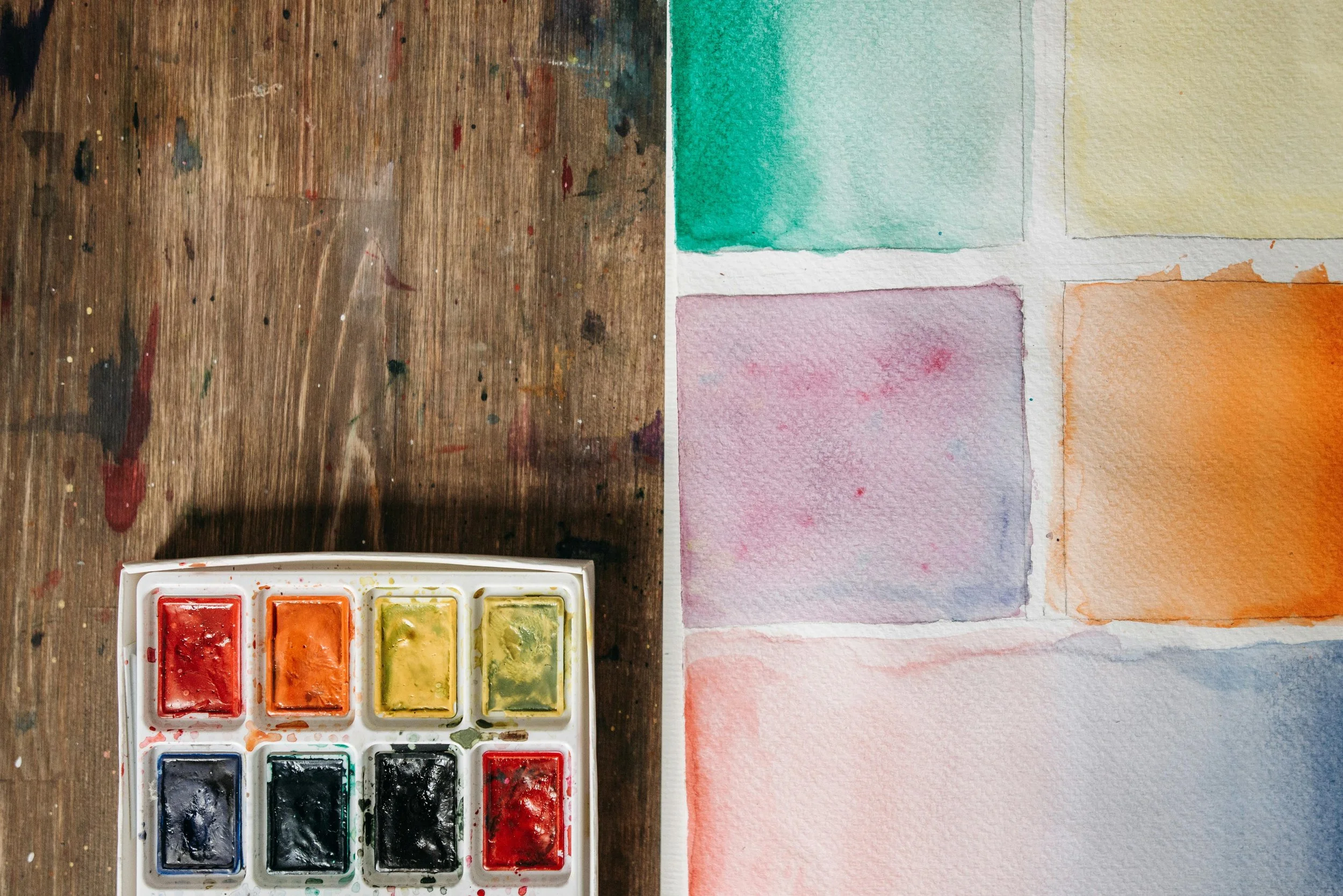 Watercolor paint set with eight colors on a wooden surface and five swatches of watercolor paper painted in different colors.