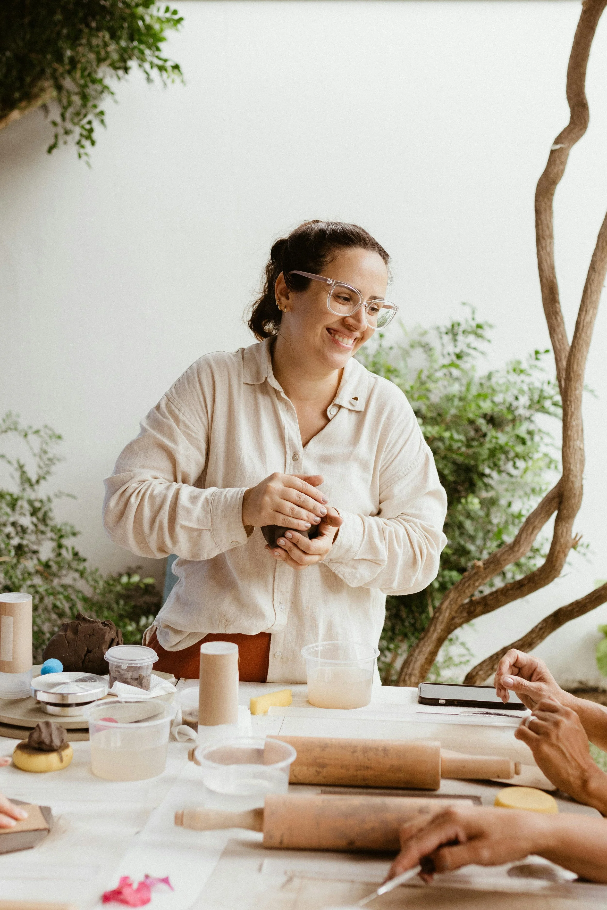 Woman smiling and holding a small bowl at a baking or cake decorating class, with various baking tools and ingredients on the table, outdoors with trees and greenery in the background.