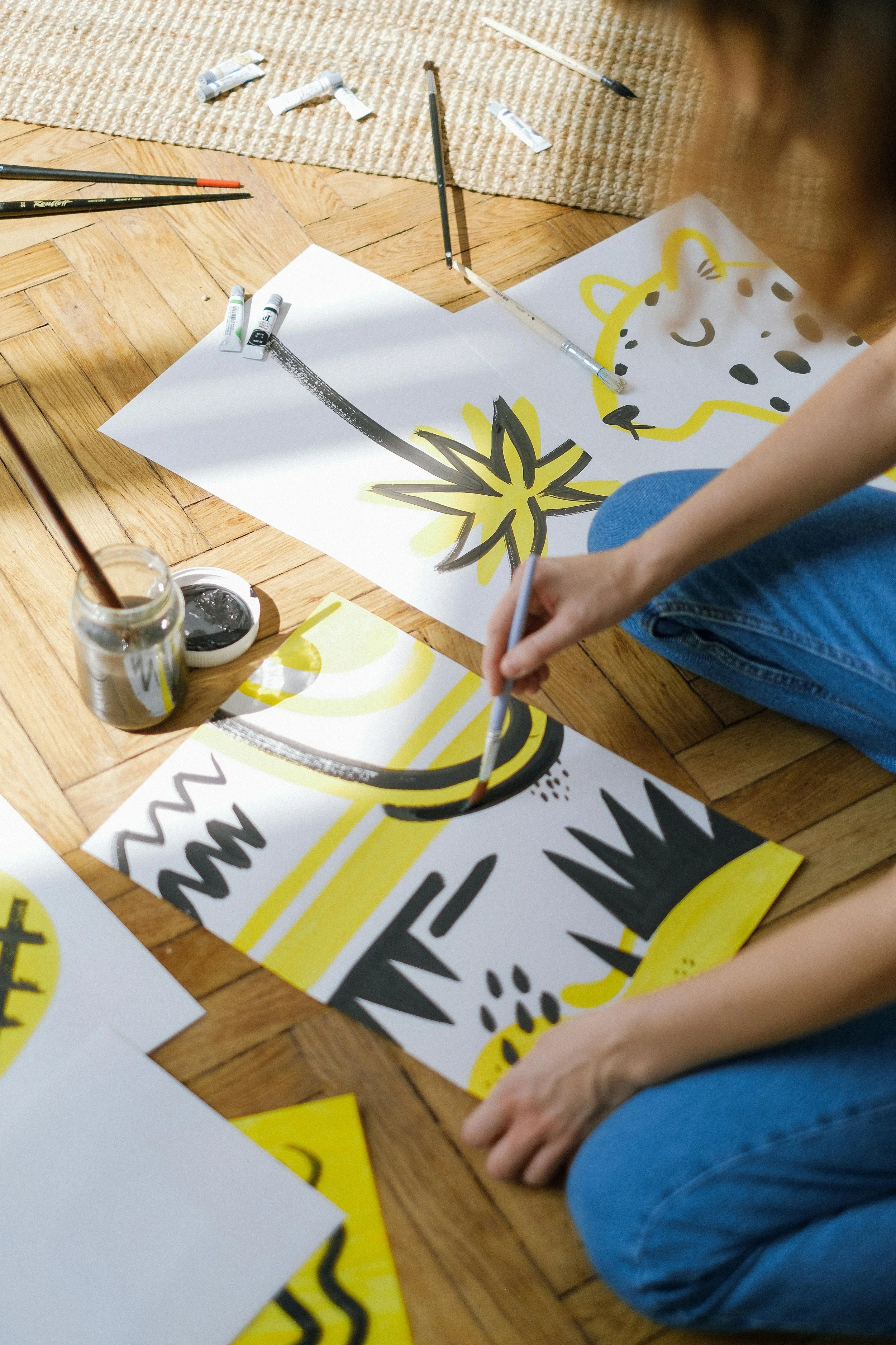 Person sitting on the floor in blue jeans creating abstract art with black, yellow, and gray paint on white paper, surrounded by paintbrushes, paint tubes, and jars of black paint.