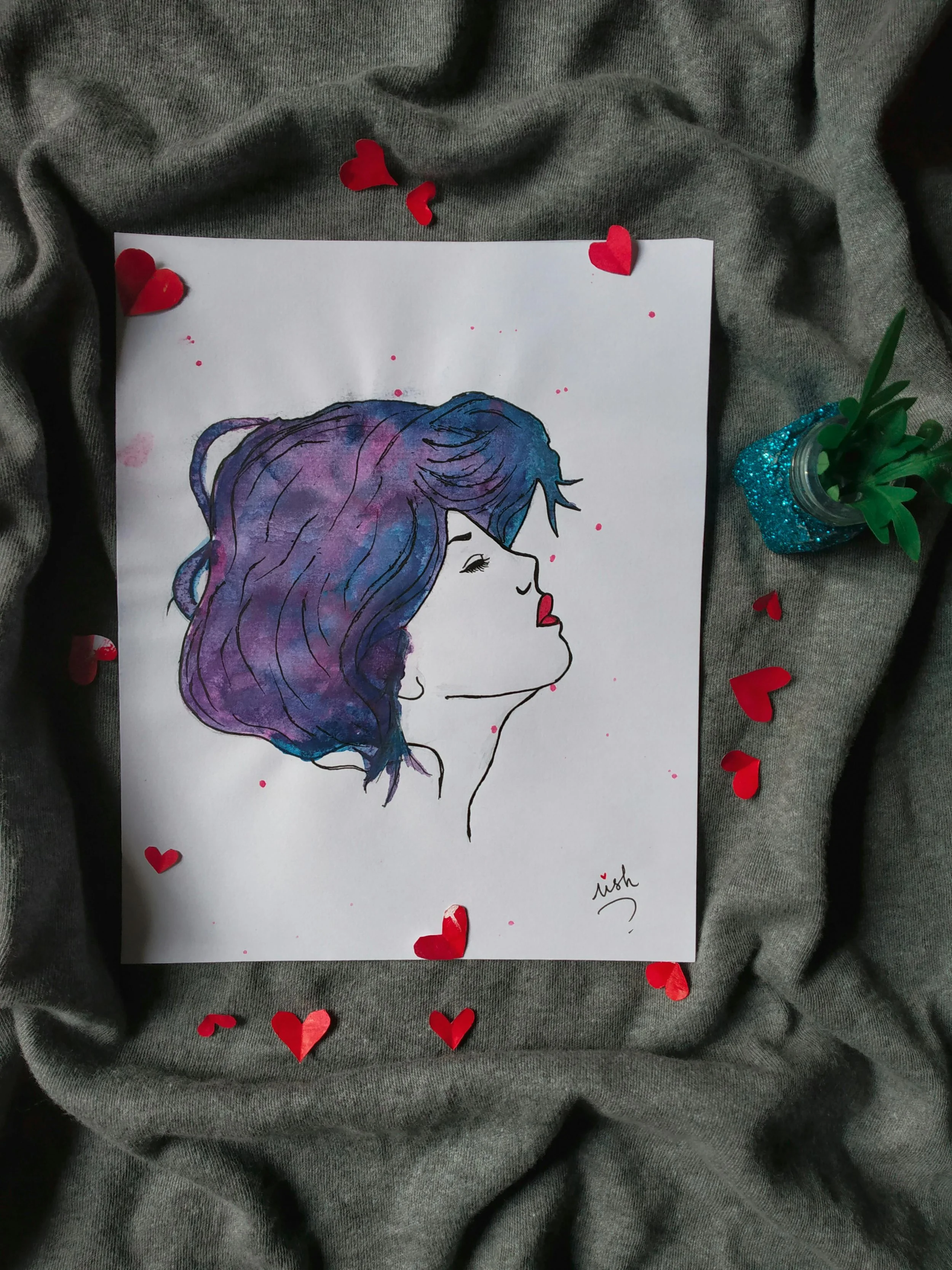 A watercolor illustration of a woman with her eyes closed, highlighting her profile. She has medium-length, wavy hair dyed in shades of purple and blue. The drawing is signed by the artist. It is surrounded by red paper hearts and placed on a gray fa