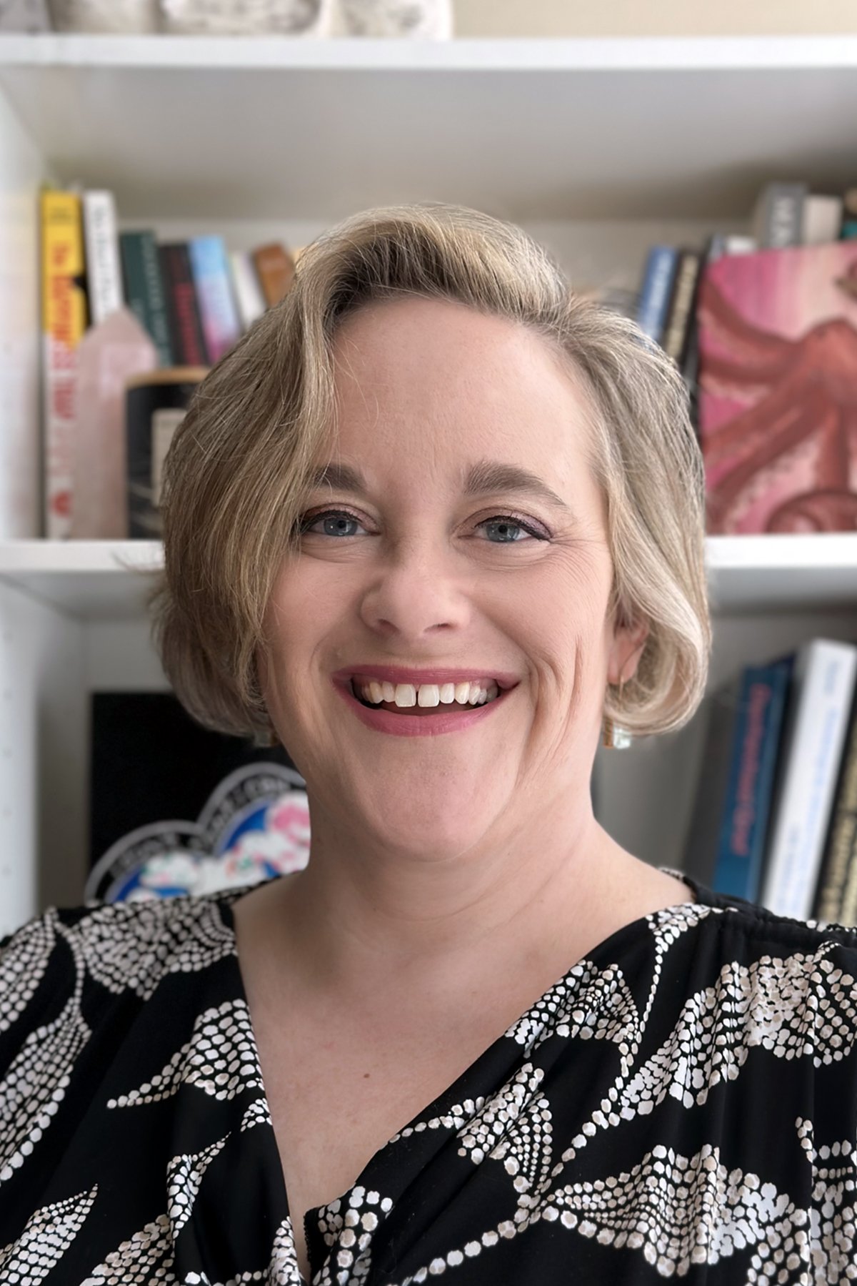 A smiling woman with short blonde hair taking a selfie in front of bookshelves filled with books and decor.