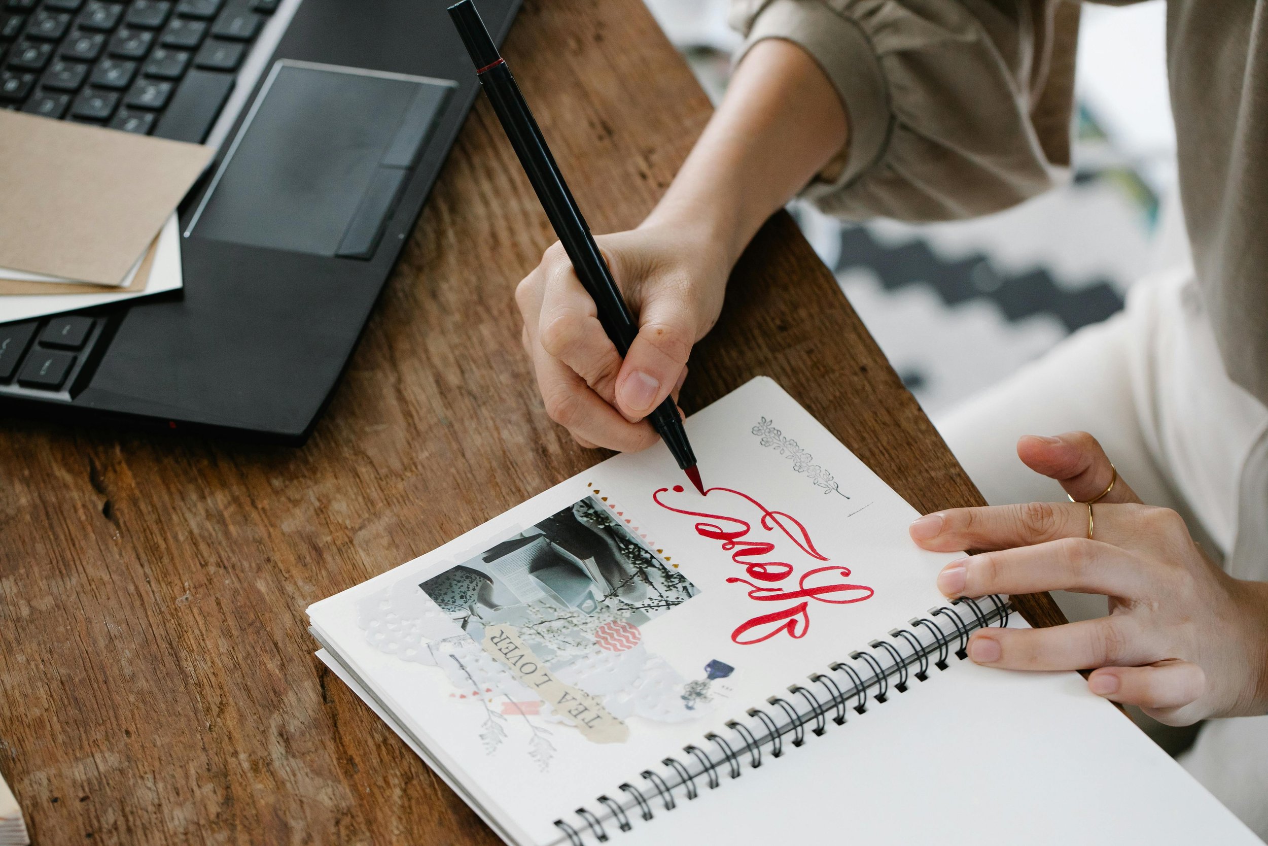 A person writing 'happy' in red cursive on a white sketchbook with decorative stickers, sitting at a wooden table next to a laptop and notebooks.