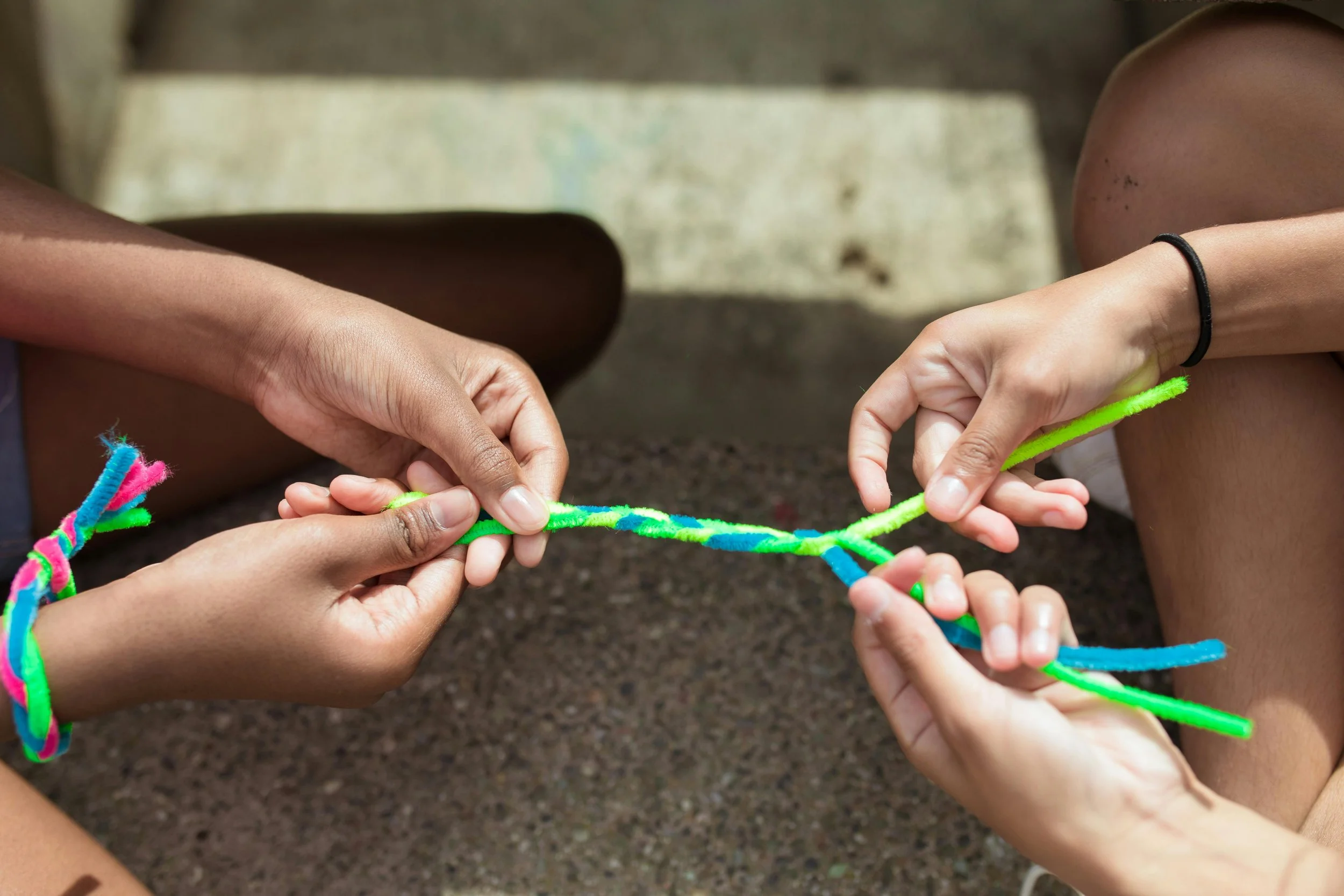 Two people are tying colorful pipe cleaners together on a rough outdoor surface.