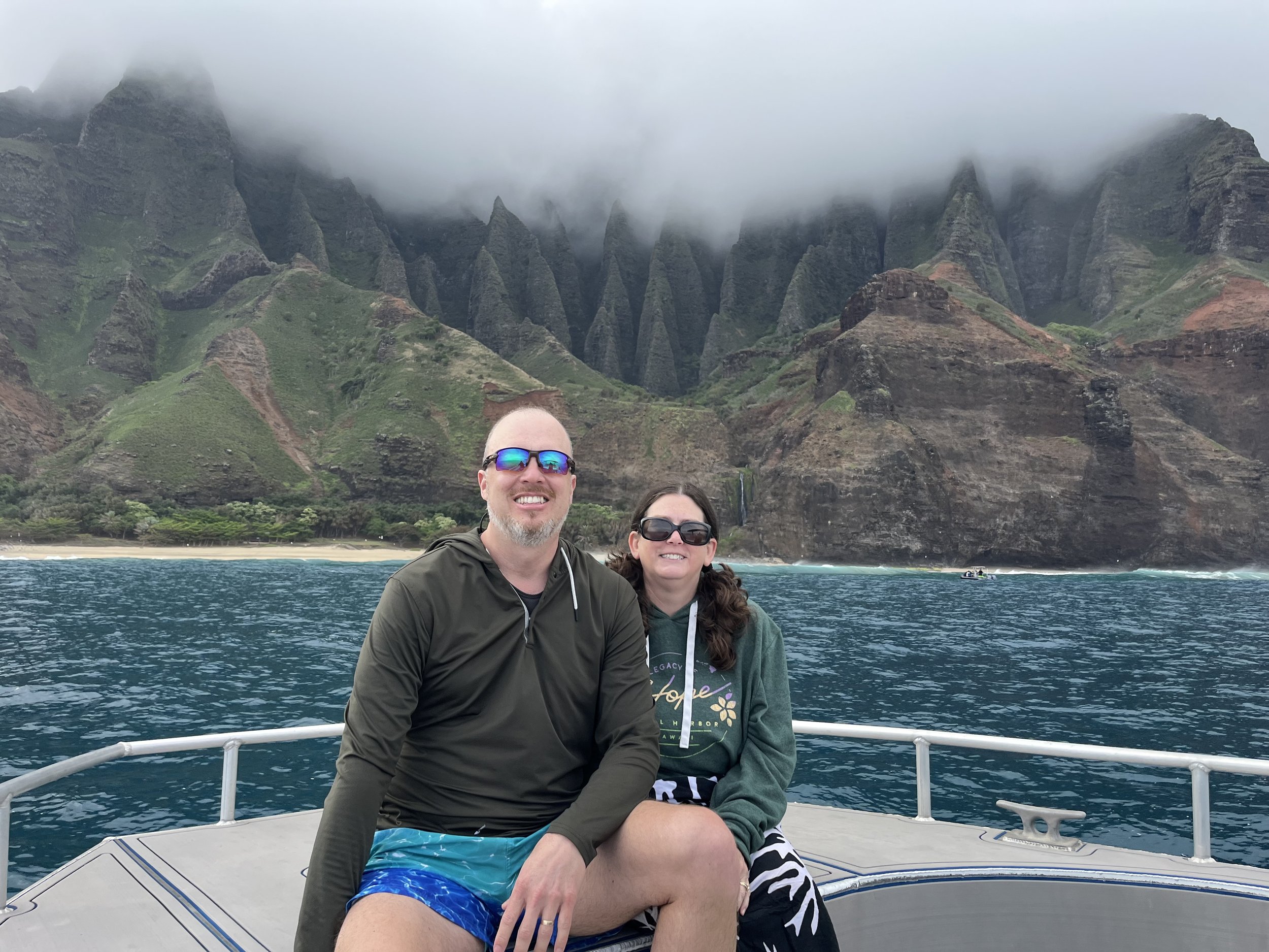 Two people sitting on the bow of a boat with green mountains and clouds in the background in a tropical location.