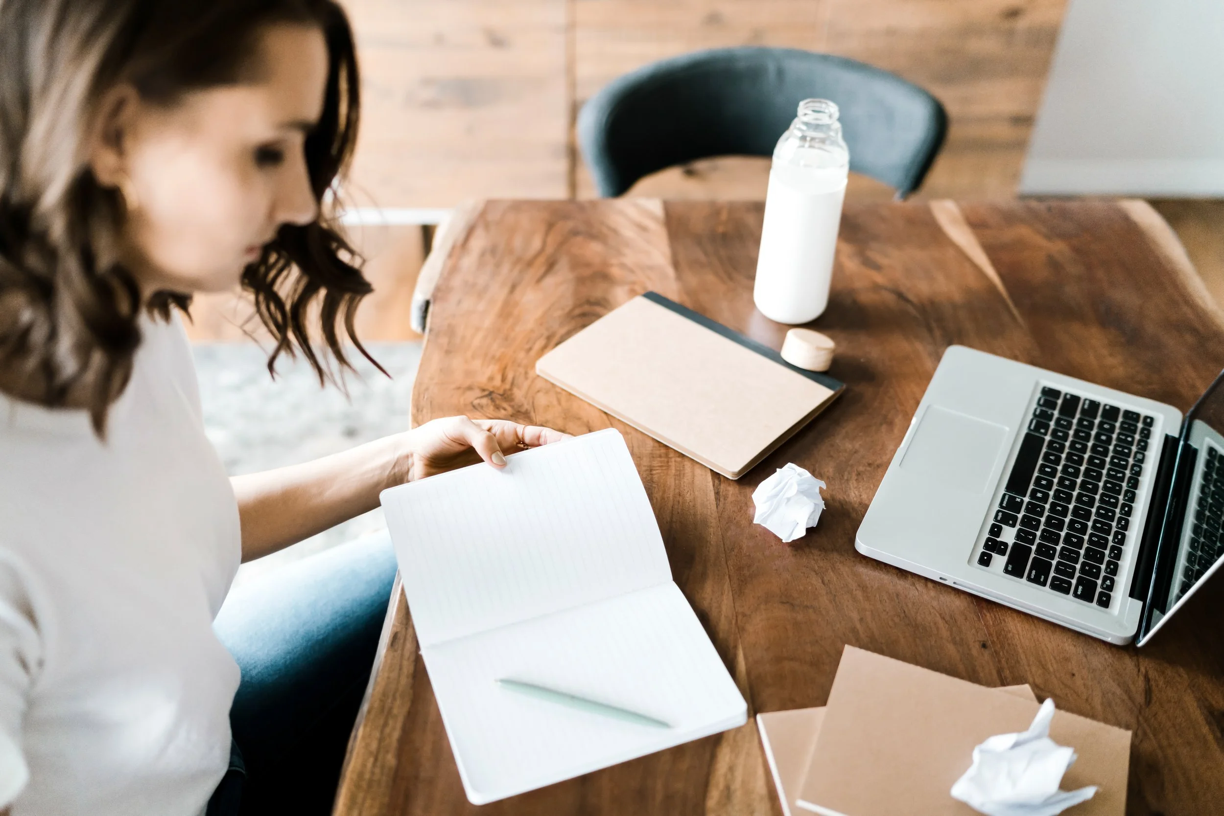 A woman sitting at a wooden table, appearing to be working or studying, with an open notebook, a laptop, crumpled tissues, a water bottle, and a closed notebook or journal on the table.
