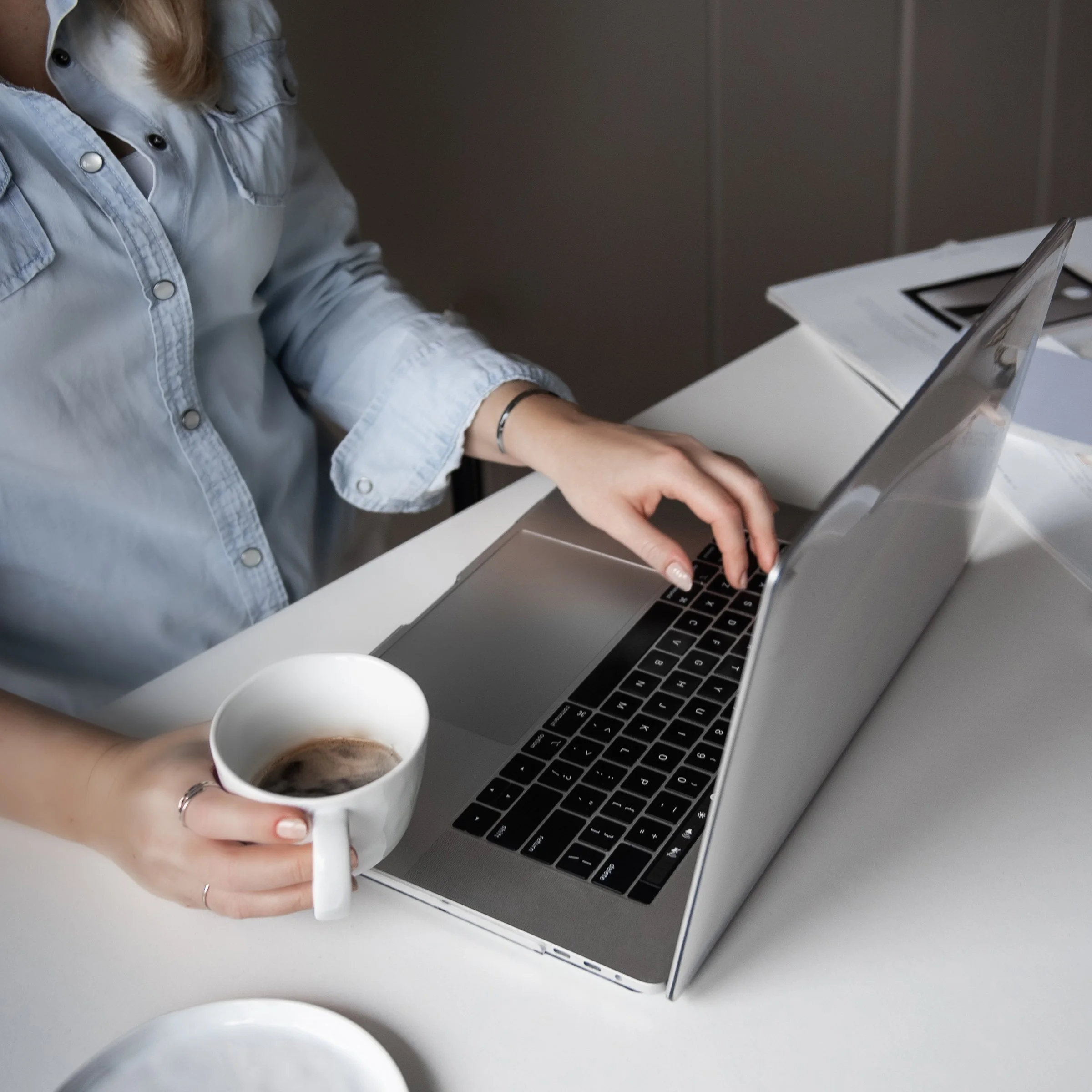 Person working on a laptop while holding a cup of coffee, sitting at a white desk with papers and a tablet.