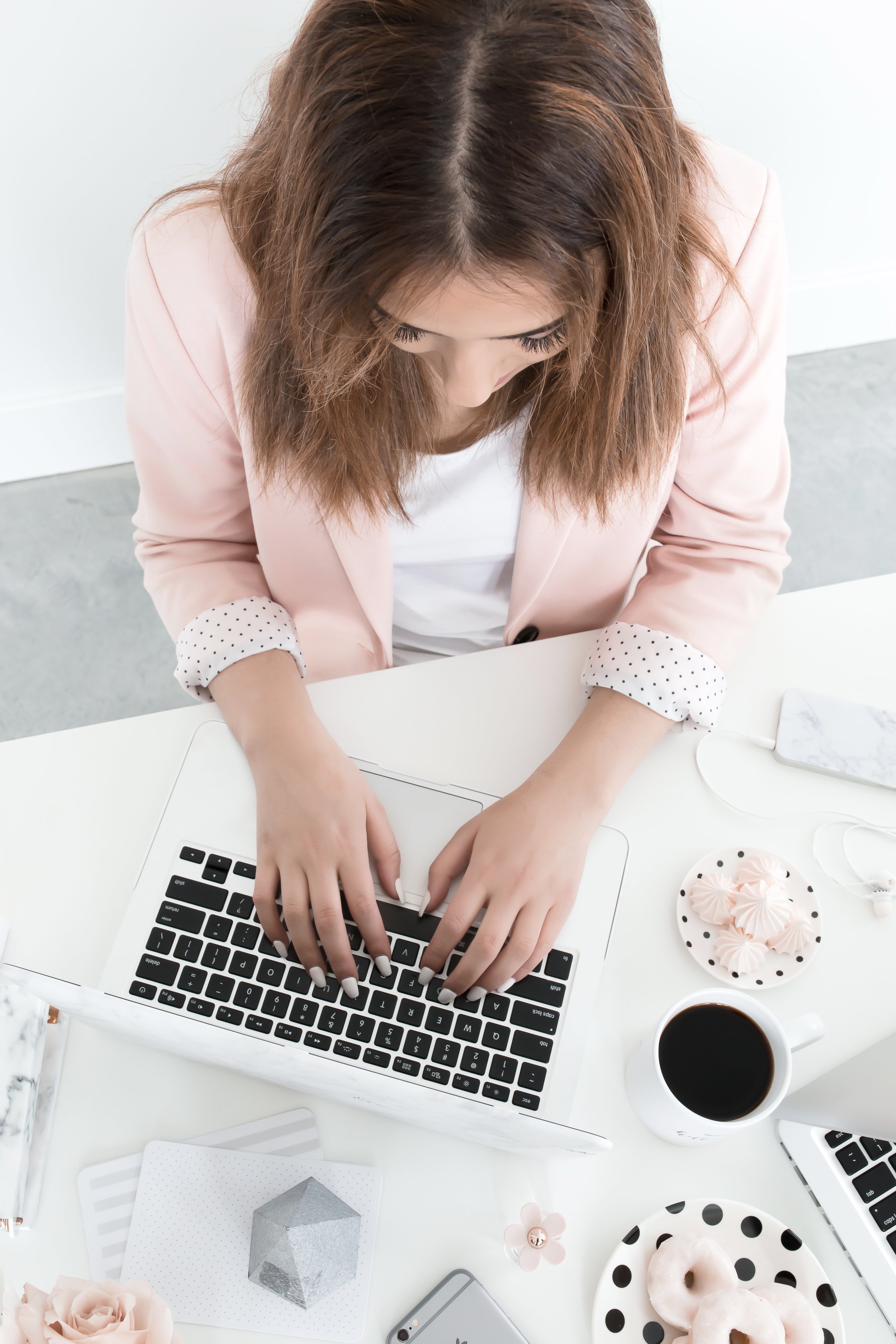 A woman with brown hair working on a laptop at a white desk surrounded by a coffee mug, decorated pastries, a phone, and various decorative objects.