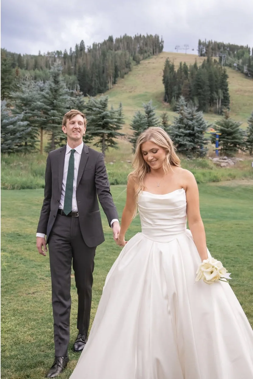 A bride in a white wedding gown holding a bouquet of white flowers, smiling, and a groom in a dark suit with a green tie, walking hand in hand outdoors on a grassy field with trees and a mountain in the background.
