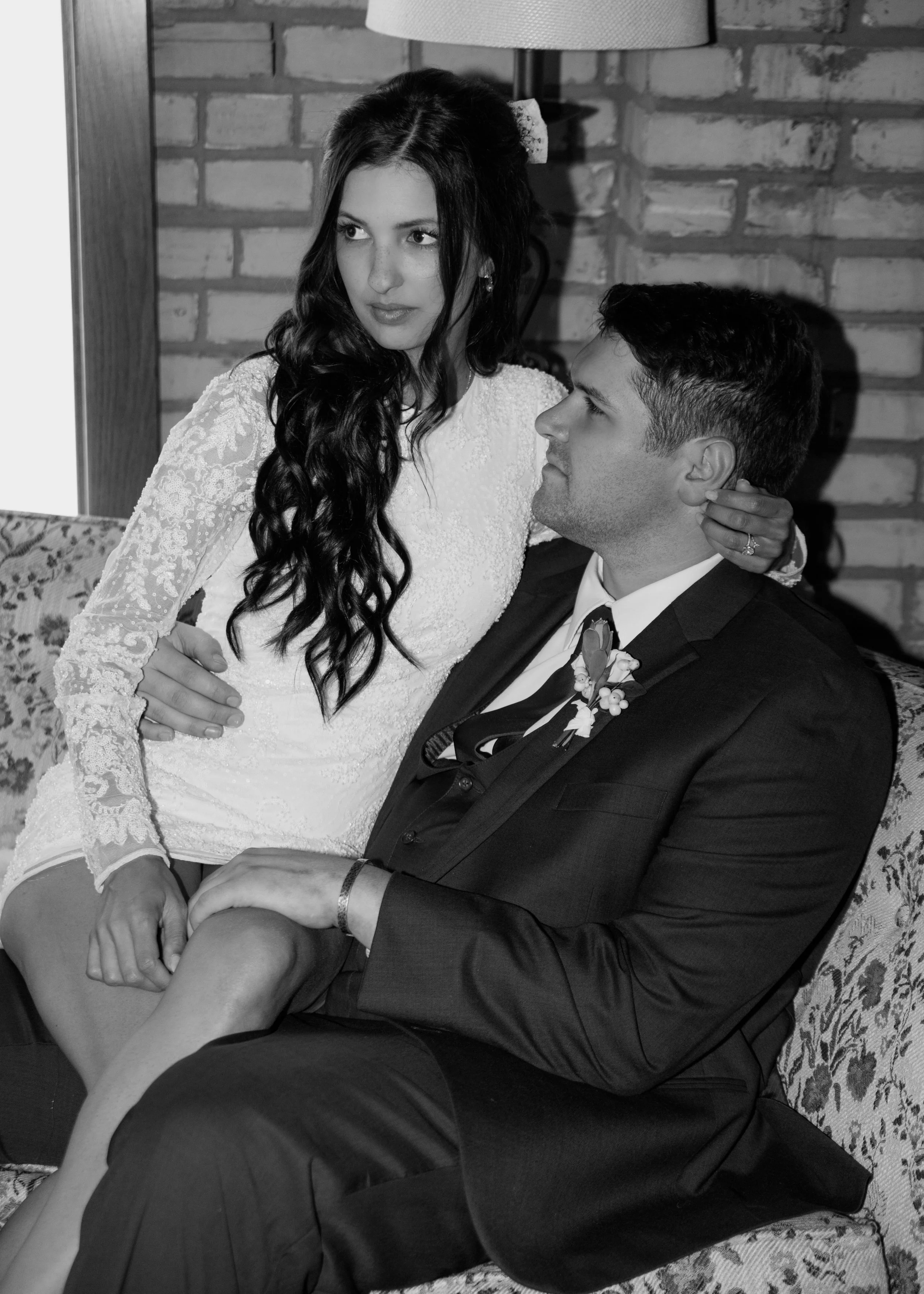 A black and white photo of a bride and groom sitting on a floral couch inside a room with a brick wall in the background, in an intimate moment.