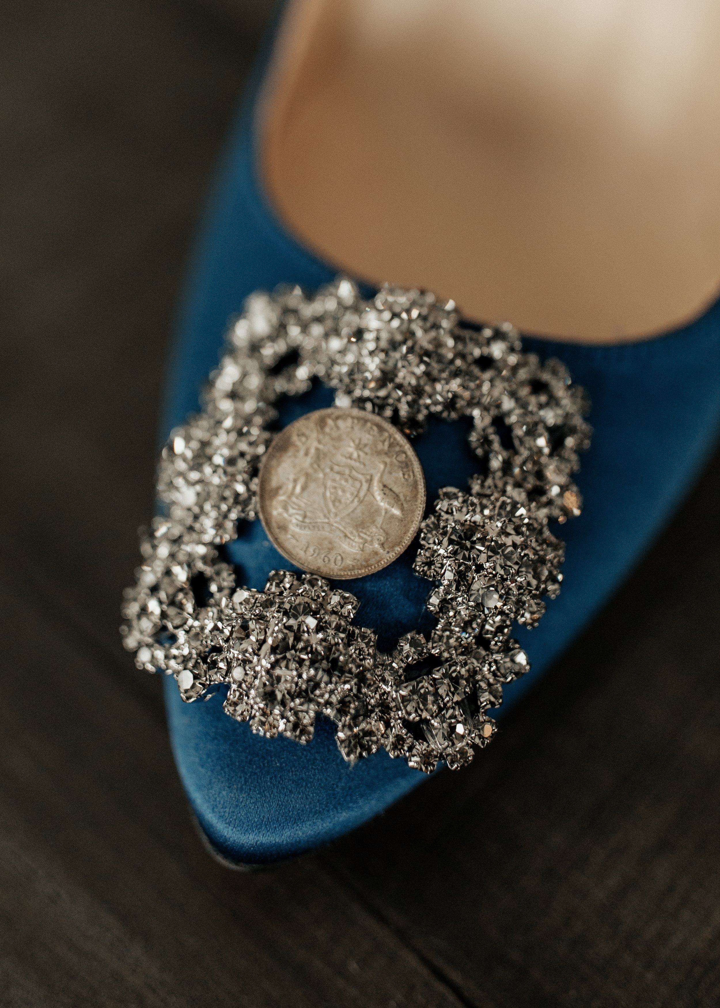 A close-up of a silver brooch with rhinestones, placed on a blue satin shoe. An old coin is on top of the brooch. The shoe has a beige heel.