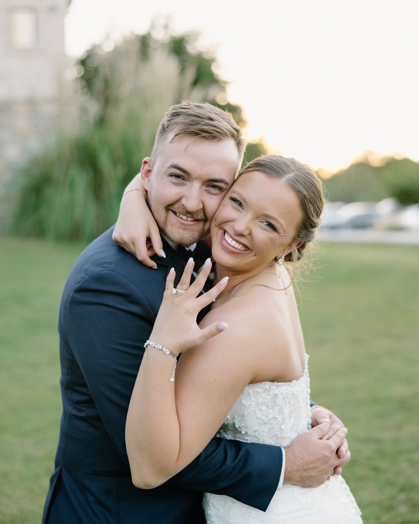 A newlywed couple embraces outdoors during sunset, smiling at the camera, with the bride showing her engagement ring.