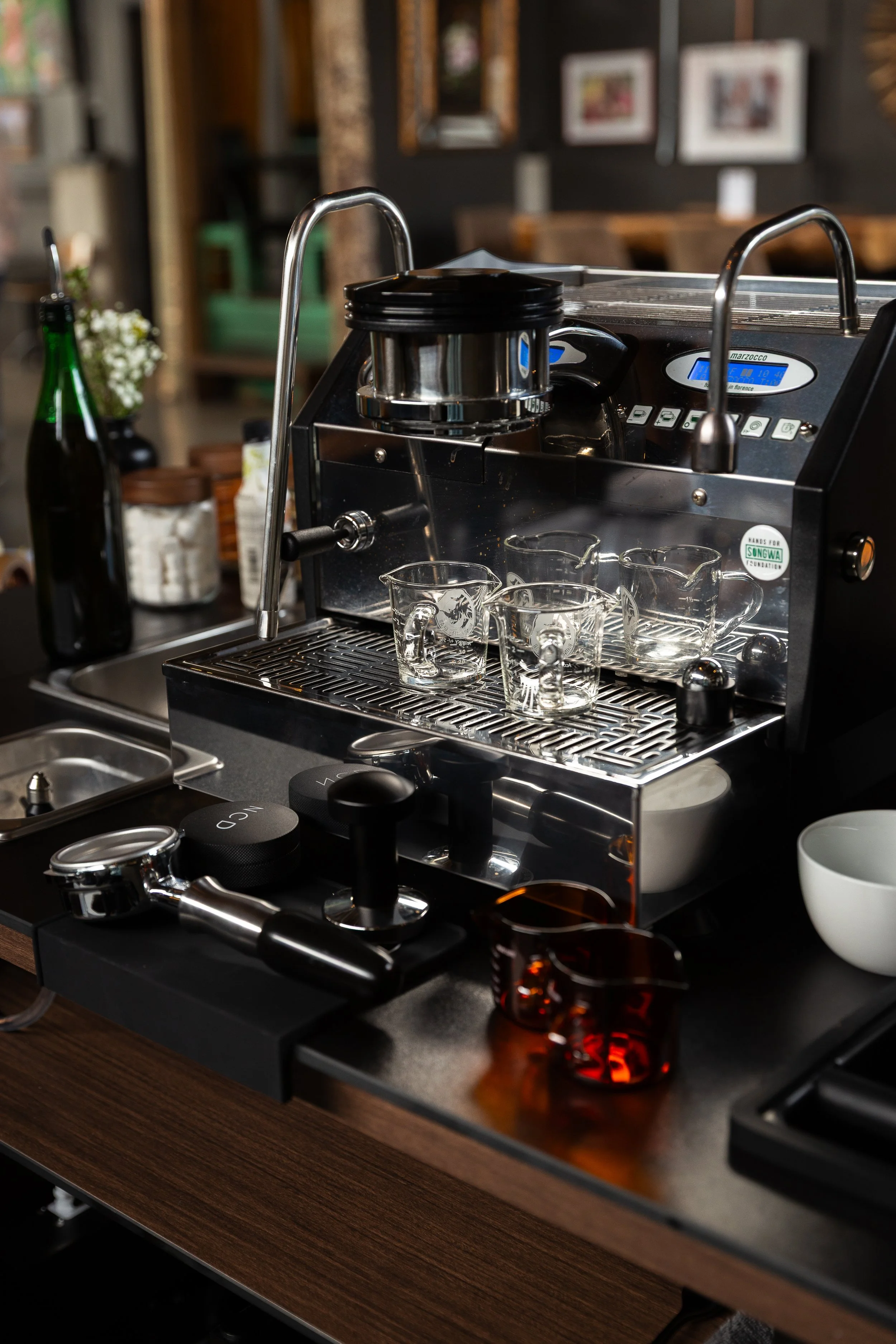 Espresso machine on a coffee shop counter with glass cups, a white bowl, a green bottle, and other kitchen items in the background.