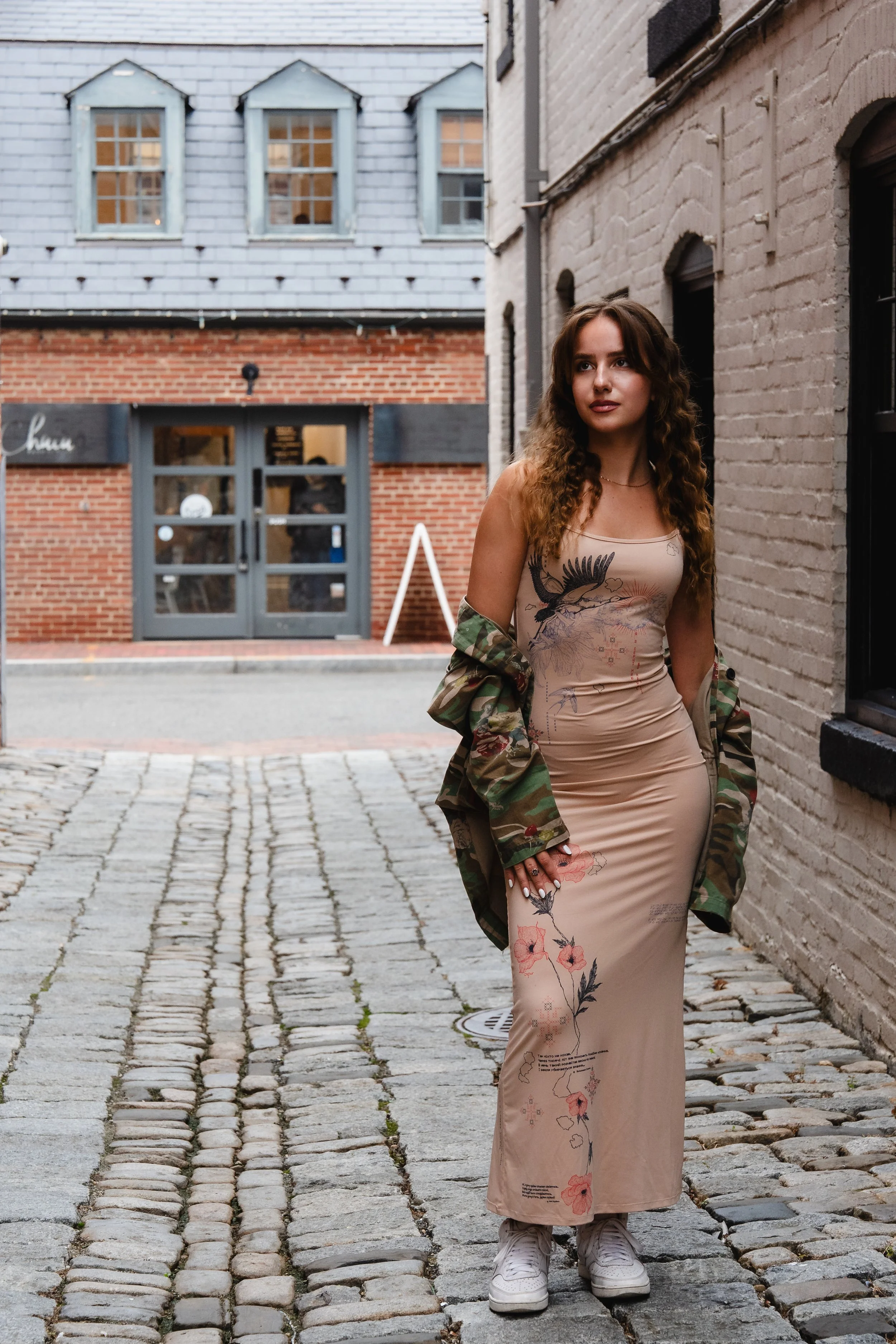 A woman with wavy hair standing on a cobblestone street between brick and stone buildings, wearing a beige floral dress, white sneakers, and a camouflage jacket draped over her shoulders.