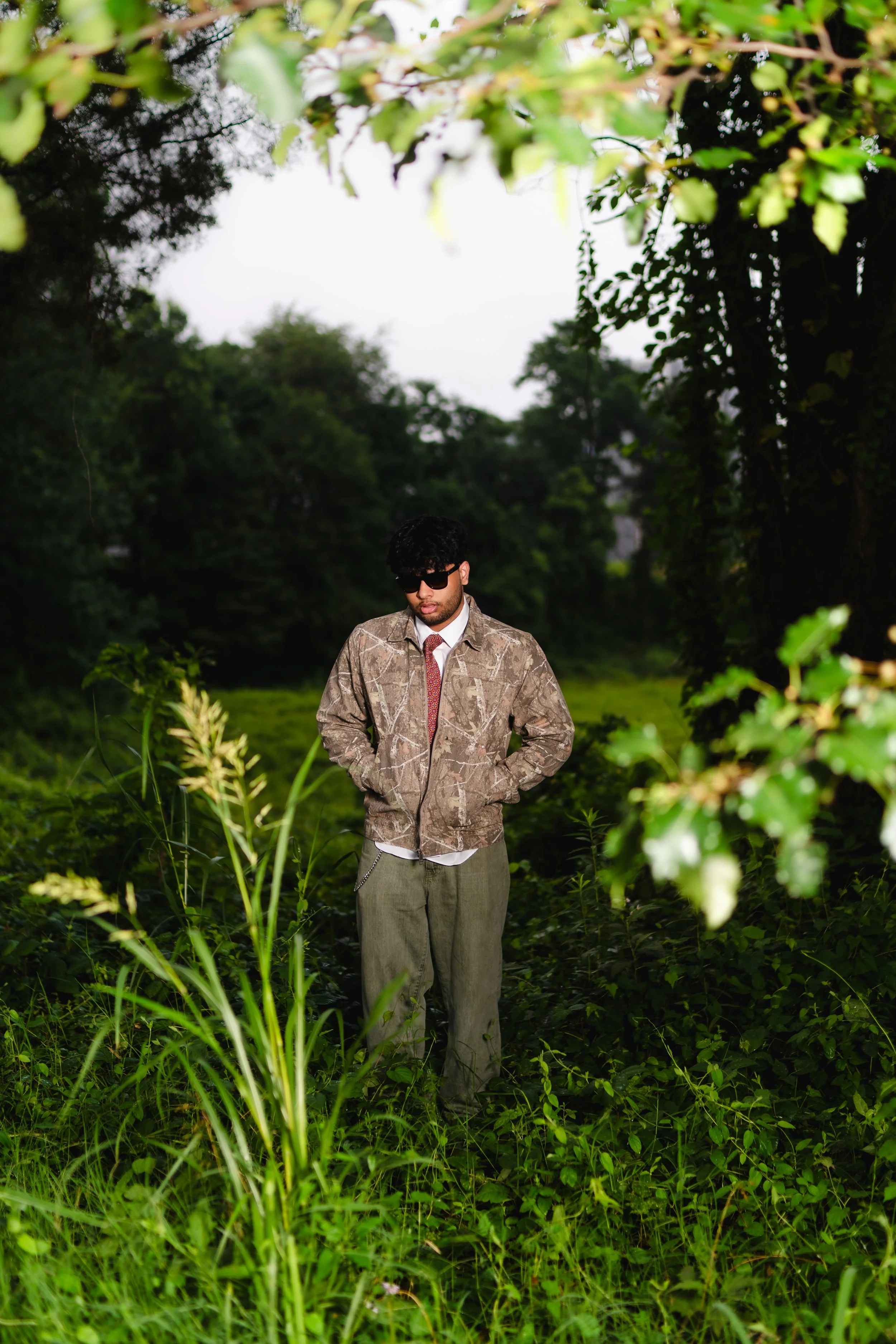 A man wearing sunglasses, a camouflage jacket, dress shirt, and tie standing in a lush green outdoor setting with trees and grass.