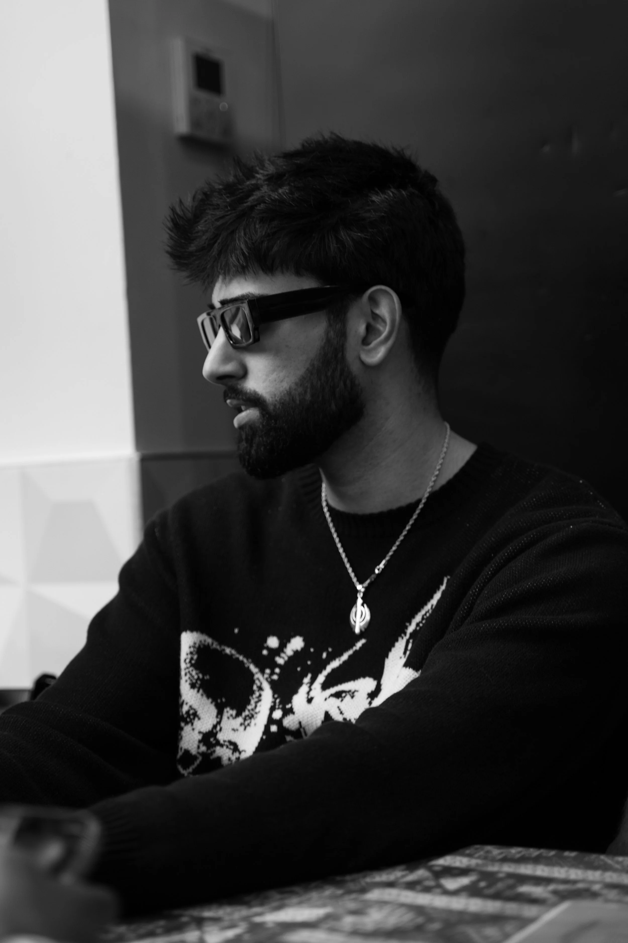 Black and white photo of a man with a beard and dark hair wearing sunglasses, a patterned sweater, and a necklace, sitting at a table in profile view.