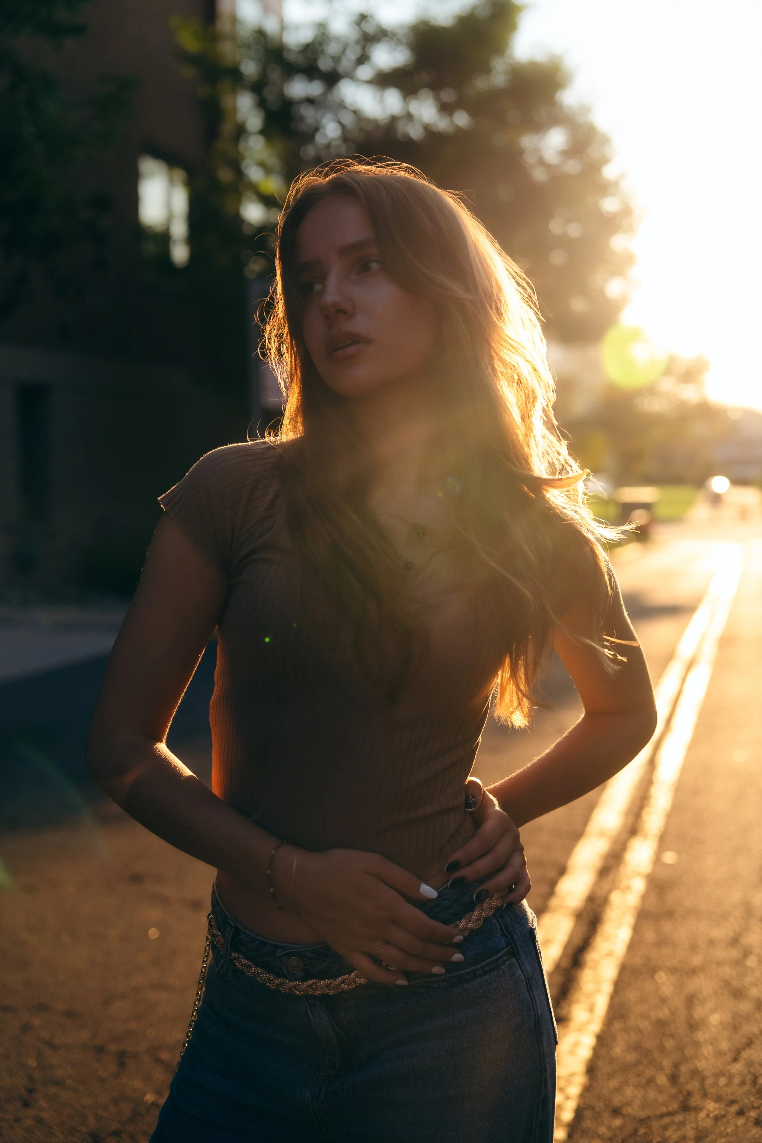 A young woman with long, wavy hair stands outdoors during sunset, with sunlight backlighting her and creating a warm glow around her.