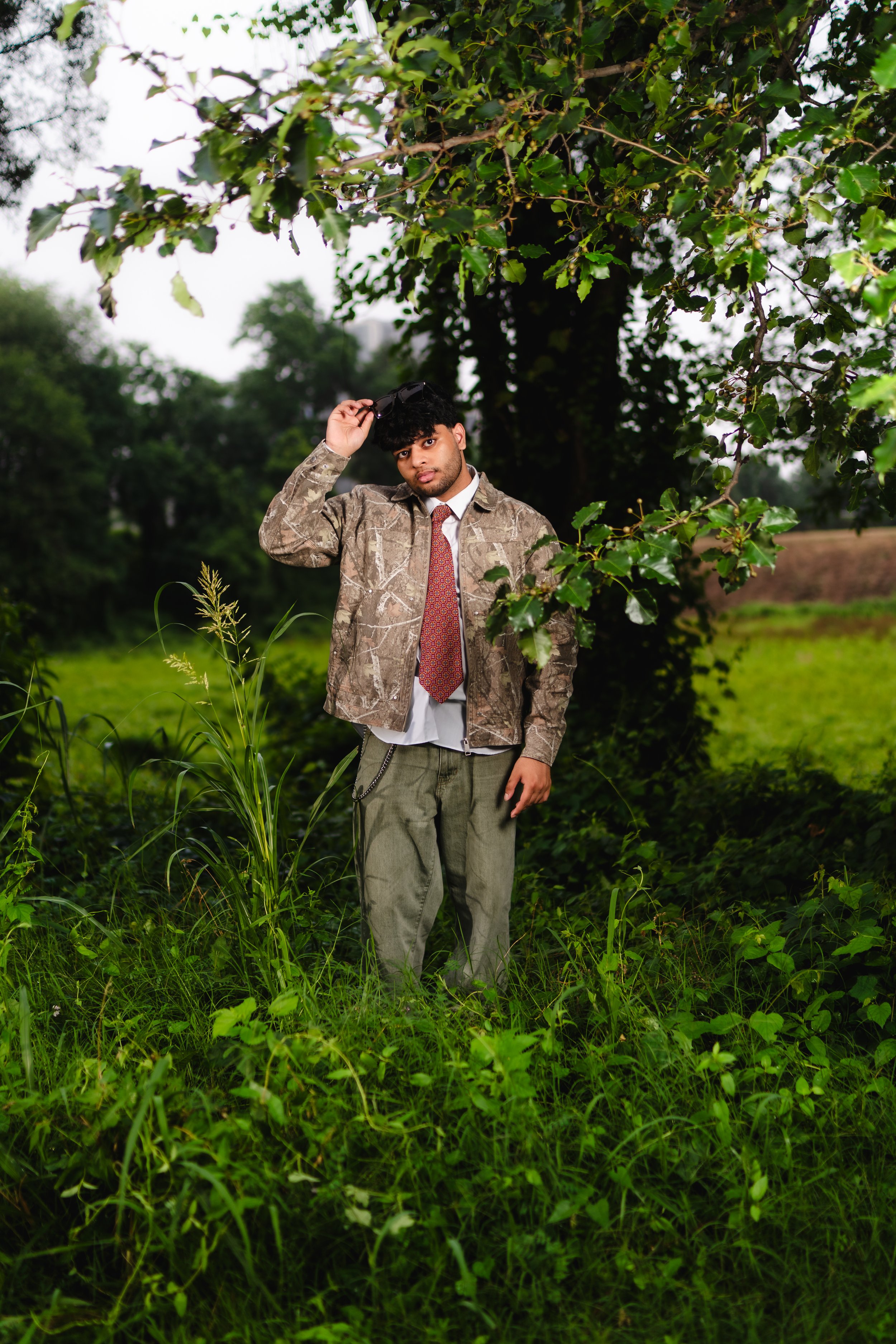 Young man dressed casually with a jacket and tie, standing outdoors in a lush green setting, leaning against a tree, with an overcast sky.