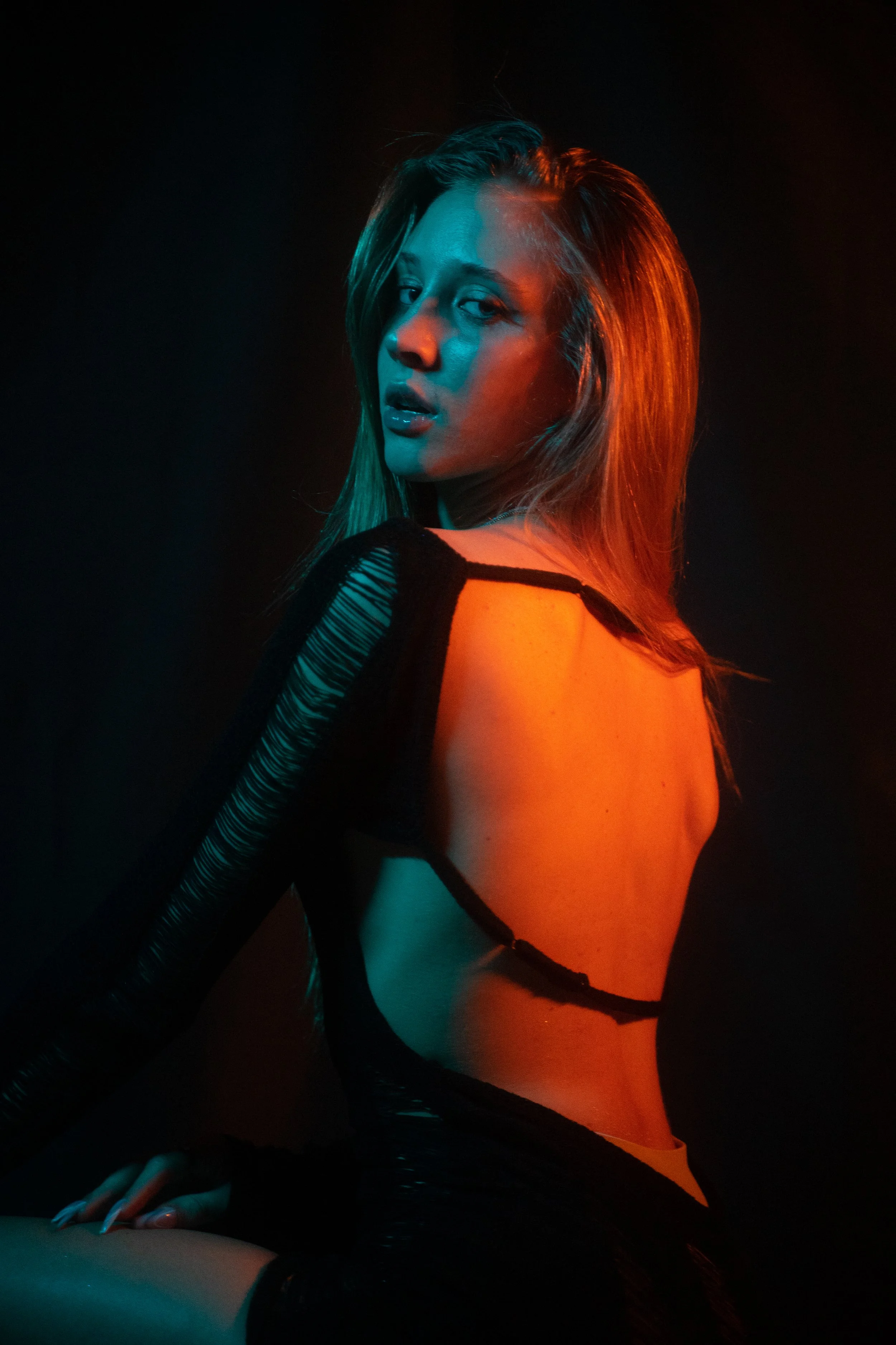 A young woman with long hair posing in a dark setting with colorful lighting, wearing a revealing black garment.
