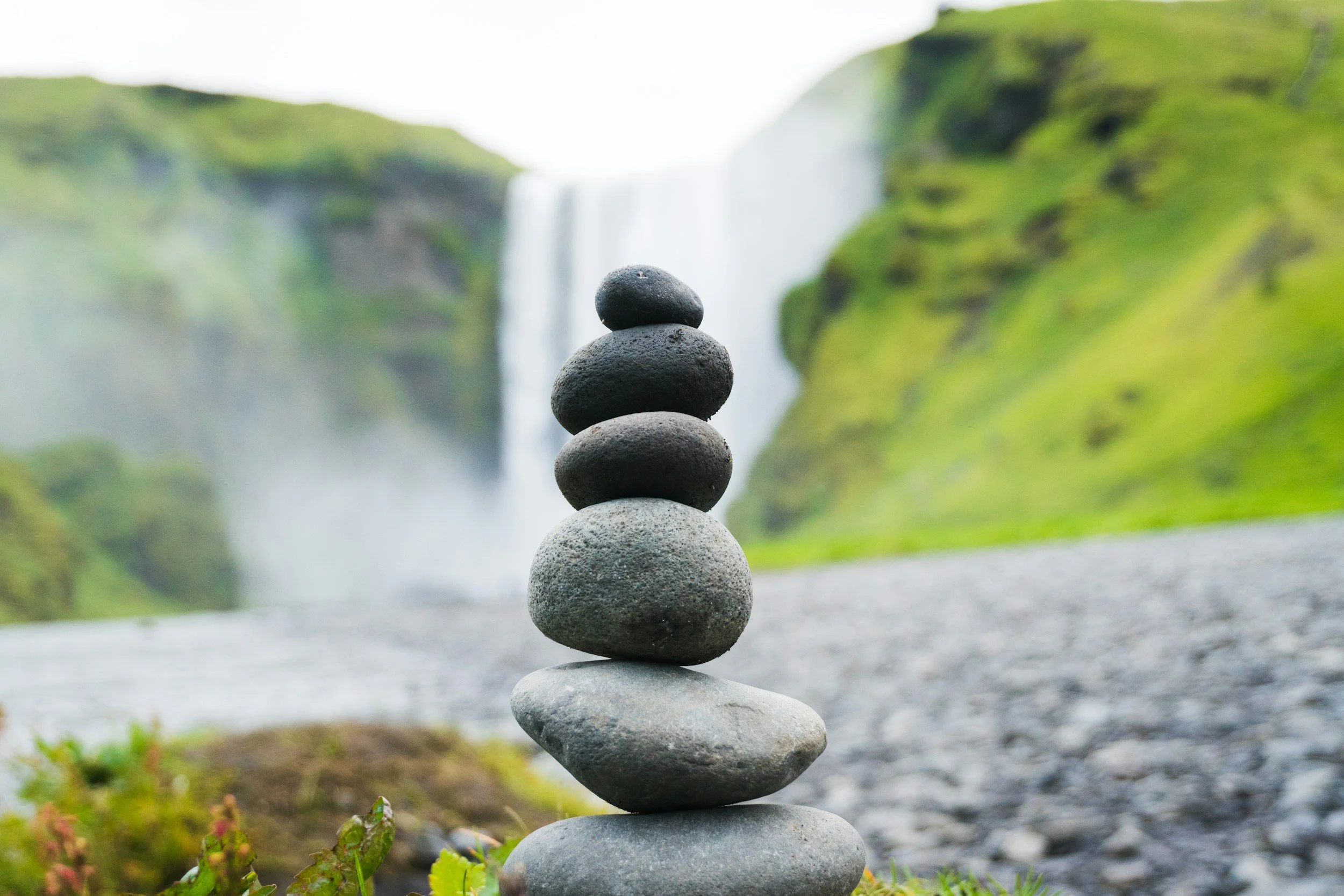Pyramid of seven stacked smooth stones in front of a waterfall and green cliffs.