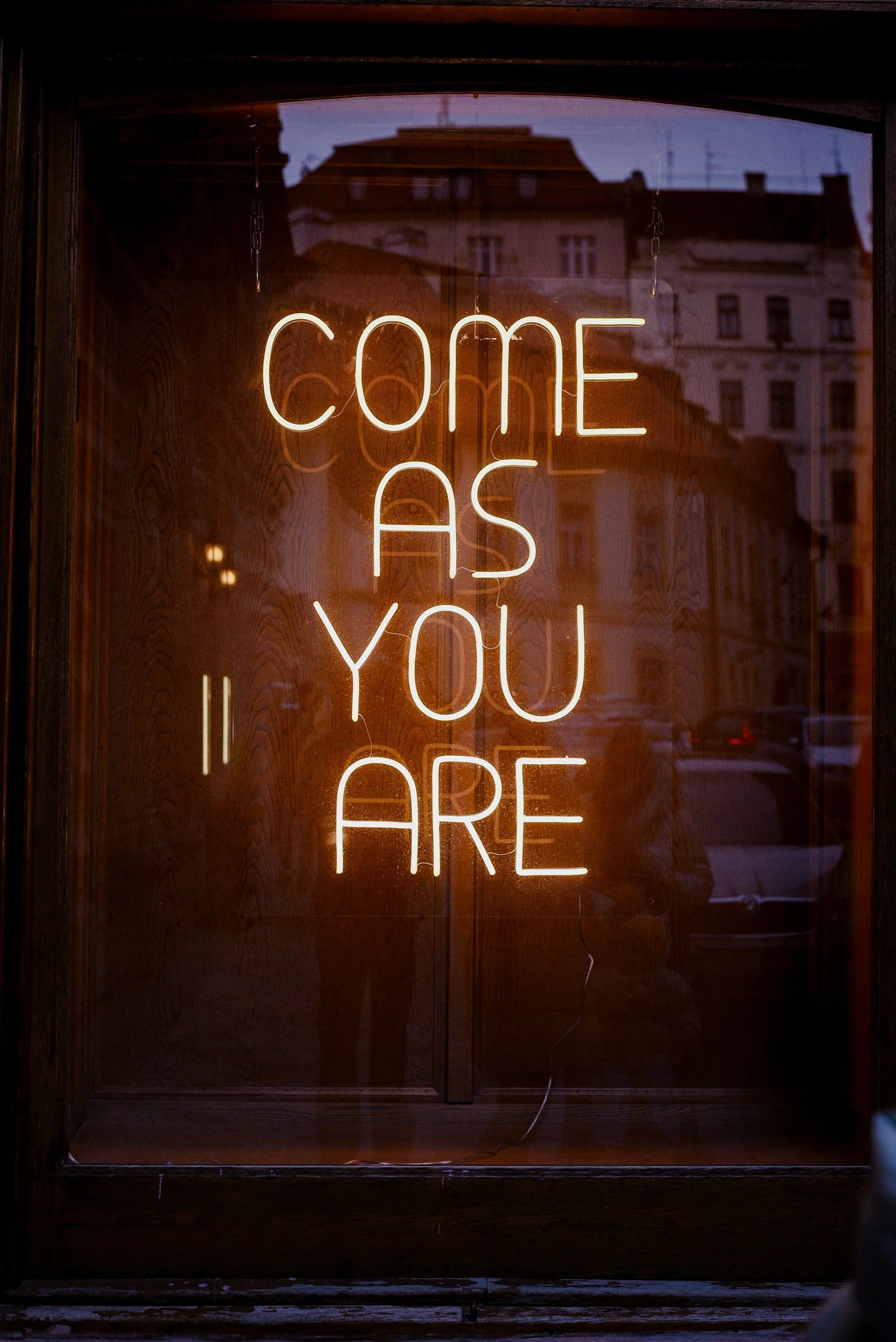 Neon sign reading 'COME AS YOU ARE' inside a window, with reflections of buildings and street scene outside.