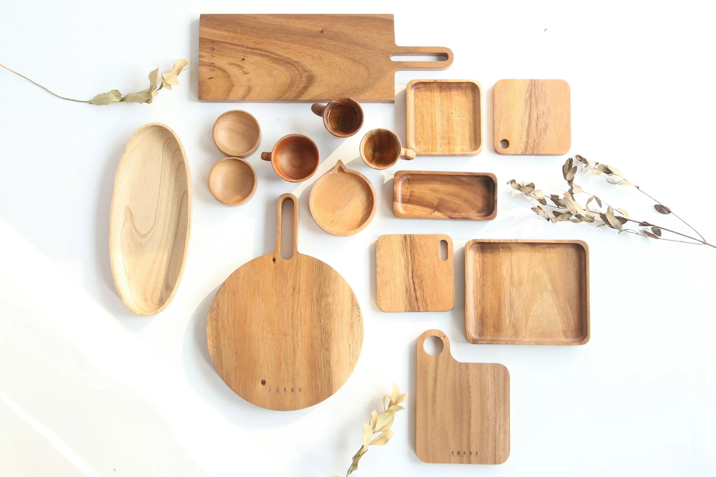 Collection of wooden kitchenware including cutting boards, bowls, trays, and small containers arranged on a white surface with dried plant decorations.