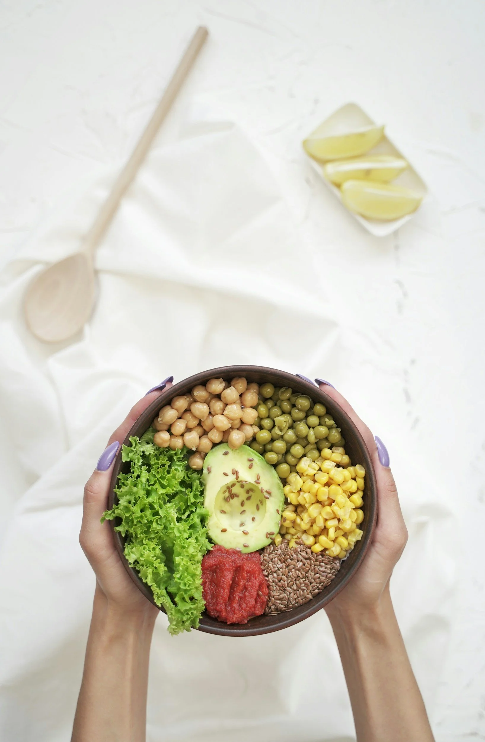 A person holding a bowl of colorful healthy food, including chickpeas, green peas, corn, lettuce, avocado, tomato, and flax seeds, with lemon wedges and a wooden spoon on a white surface in the background.