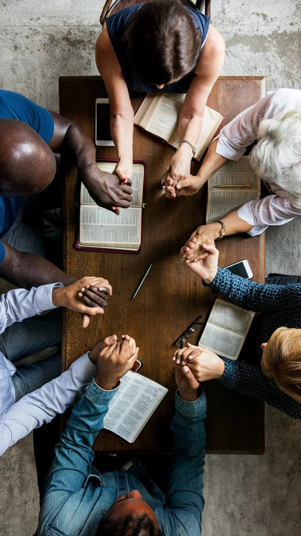 Top-down view of a diverse group of six people holding hands across a wooden table with open books, tablets, and a smartphone.