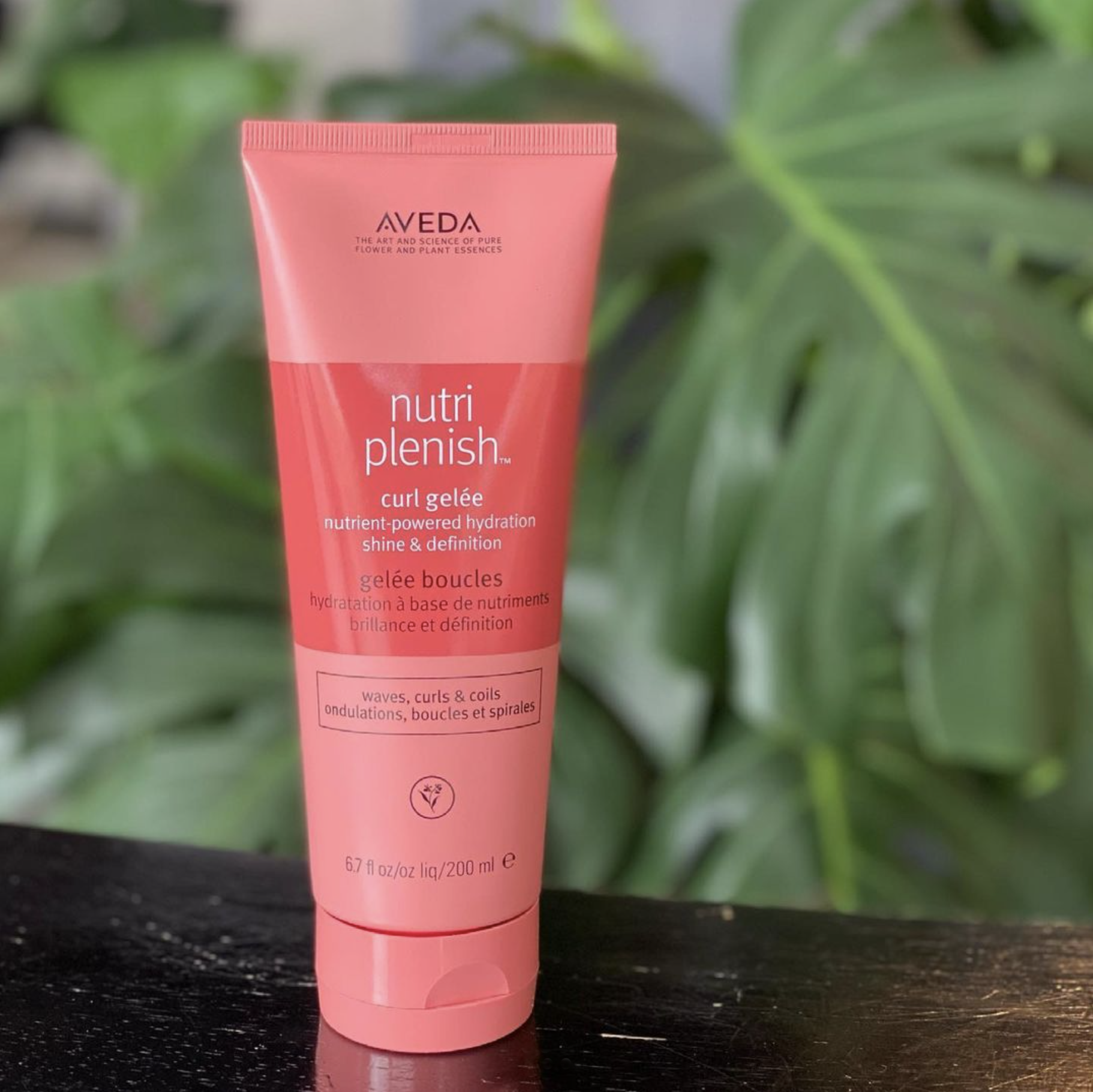 A pink tube of Aveda Nutri Plenish conditioner on a white surface, surrounded by two gold hoop earrings and pink books or boxes.