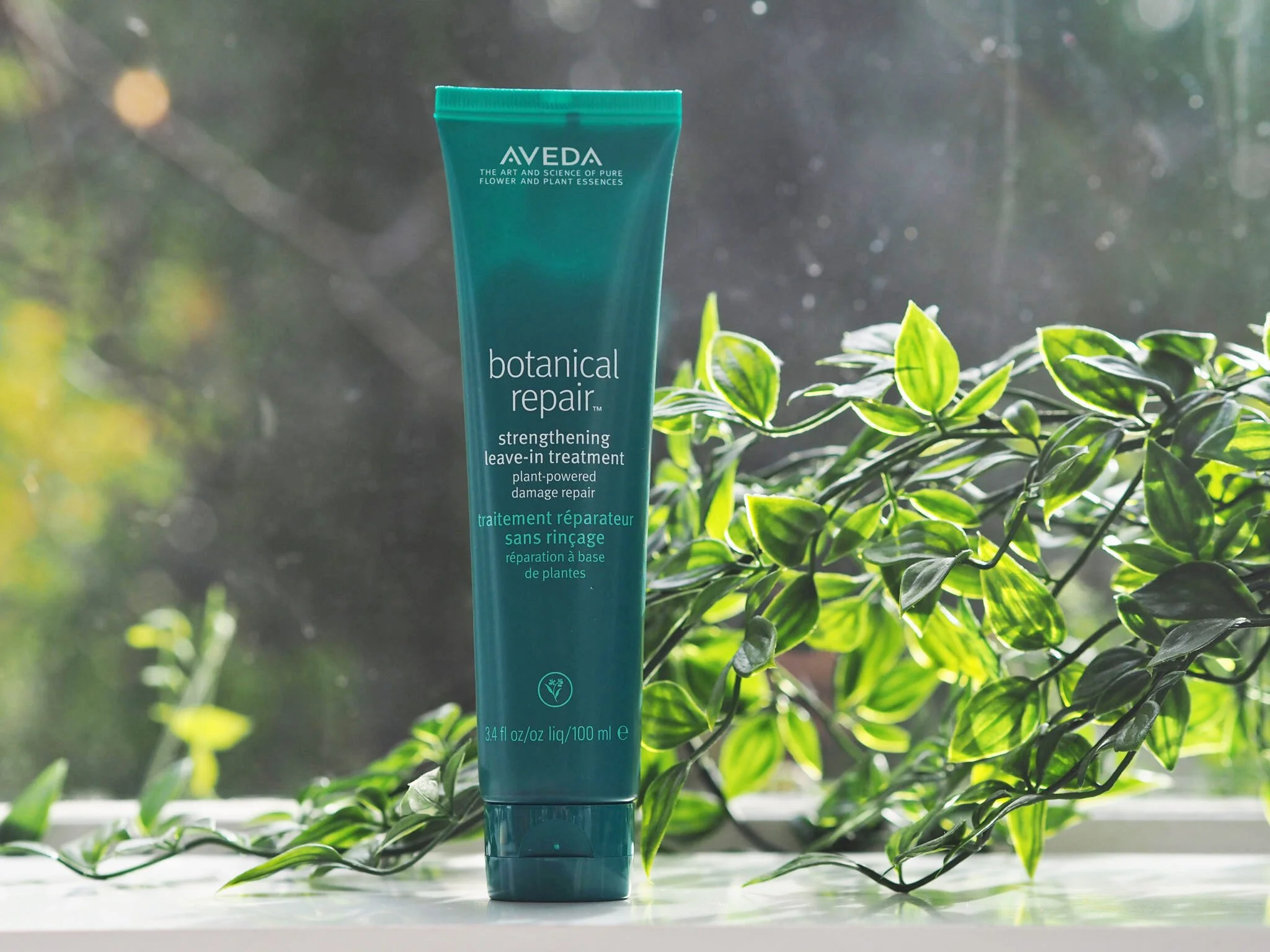 A tube of Aveda Botanical Repair strengthening leave-in treatment placed on a white surface with green plants and sunlight in the background.