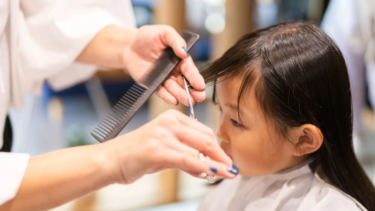 A young girl getting her hair cut at a salon, with a stylist holding scissors and a comb.